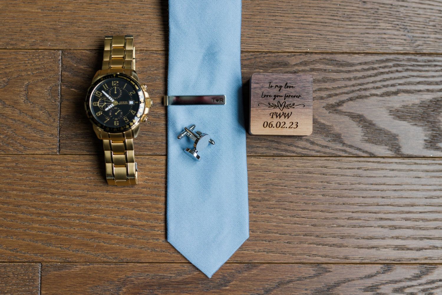 Wedding accessories including a gold watch, light blue tie, and handwritten note arranged on wooden surface.