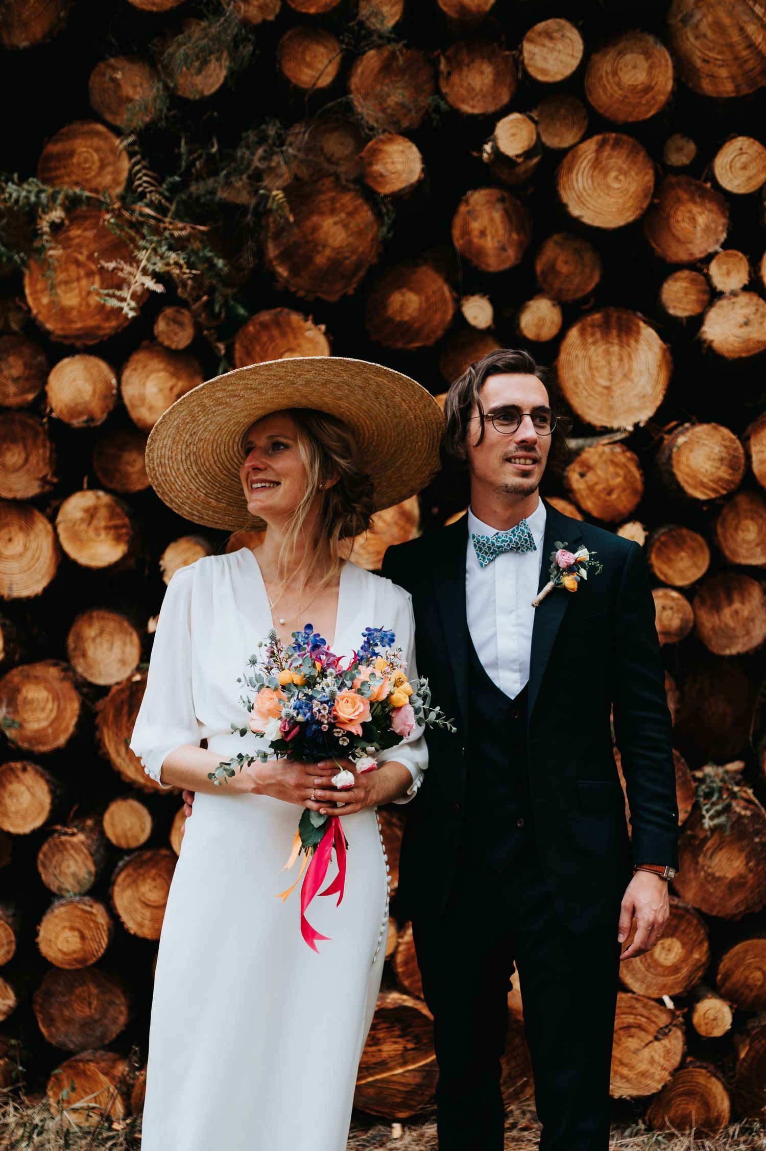 A couple in wedding attire shares a joyful moment against a wall of stacked firewood.
