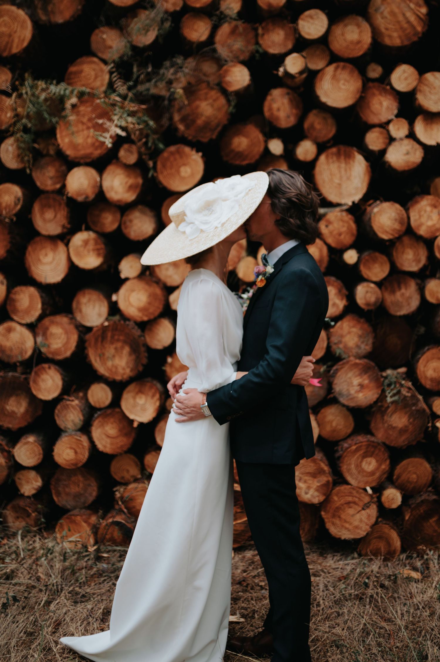 A couple poses against a backdrop of stacked logs in elegant wedding attire.