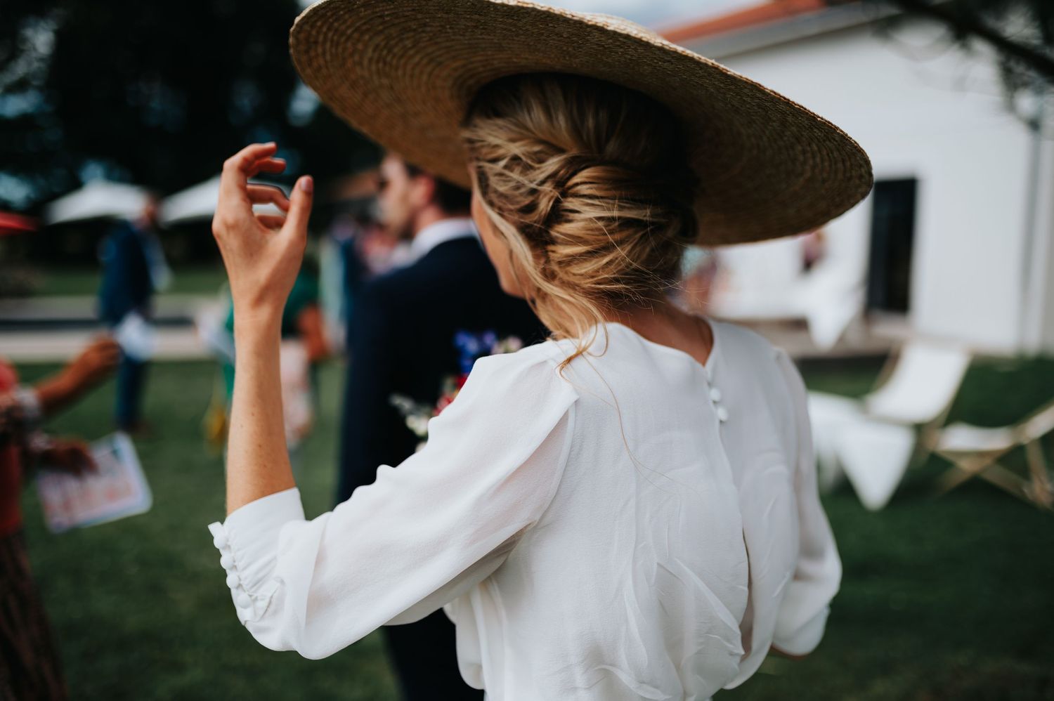 Person in white dress and wide-brimmed hat stands against woodpile background.