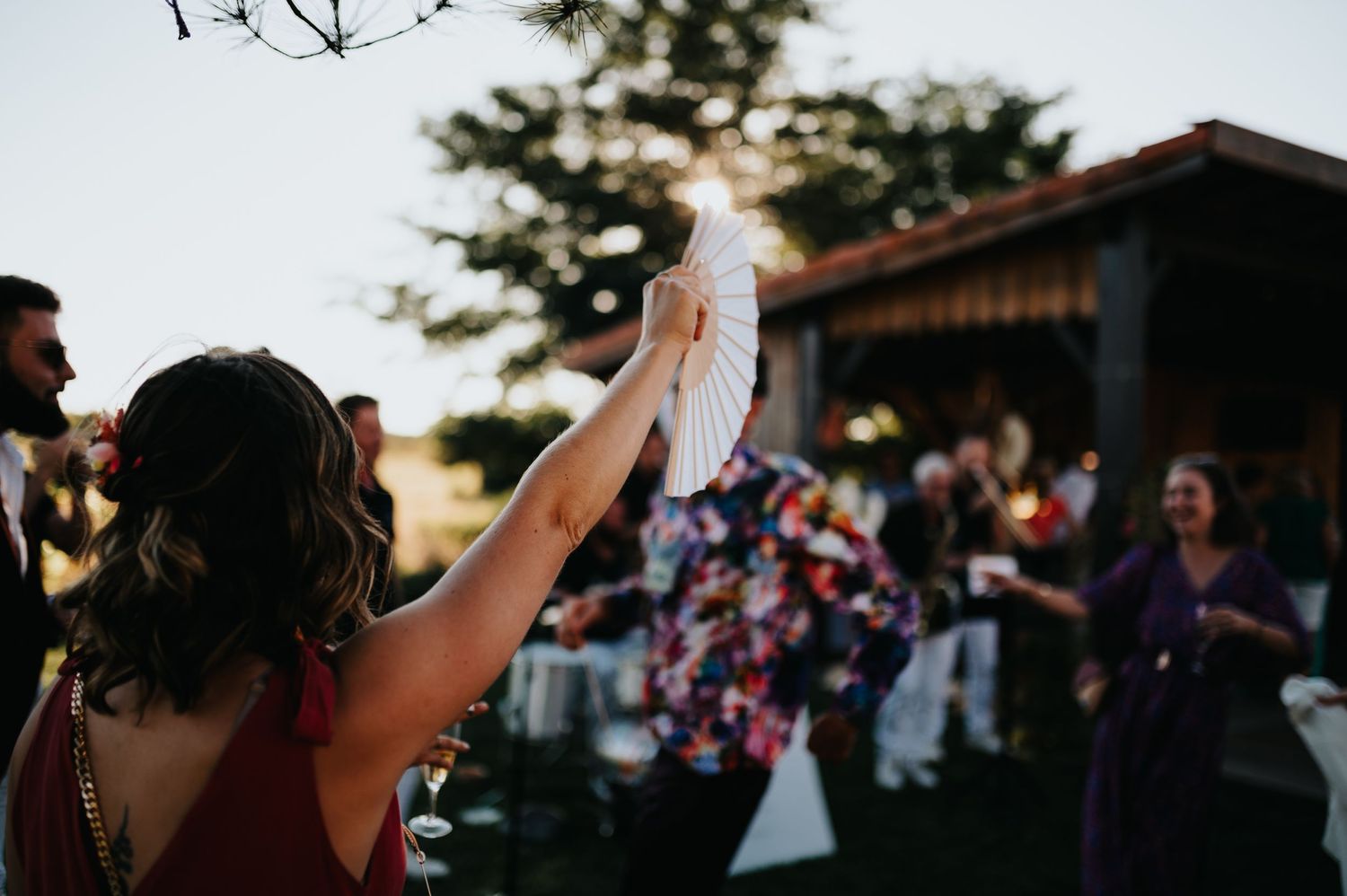 Wedding party dancing and celebrating outdoors at dusk near a wooden structure.