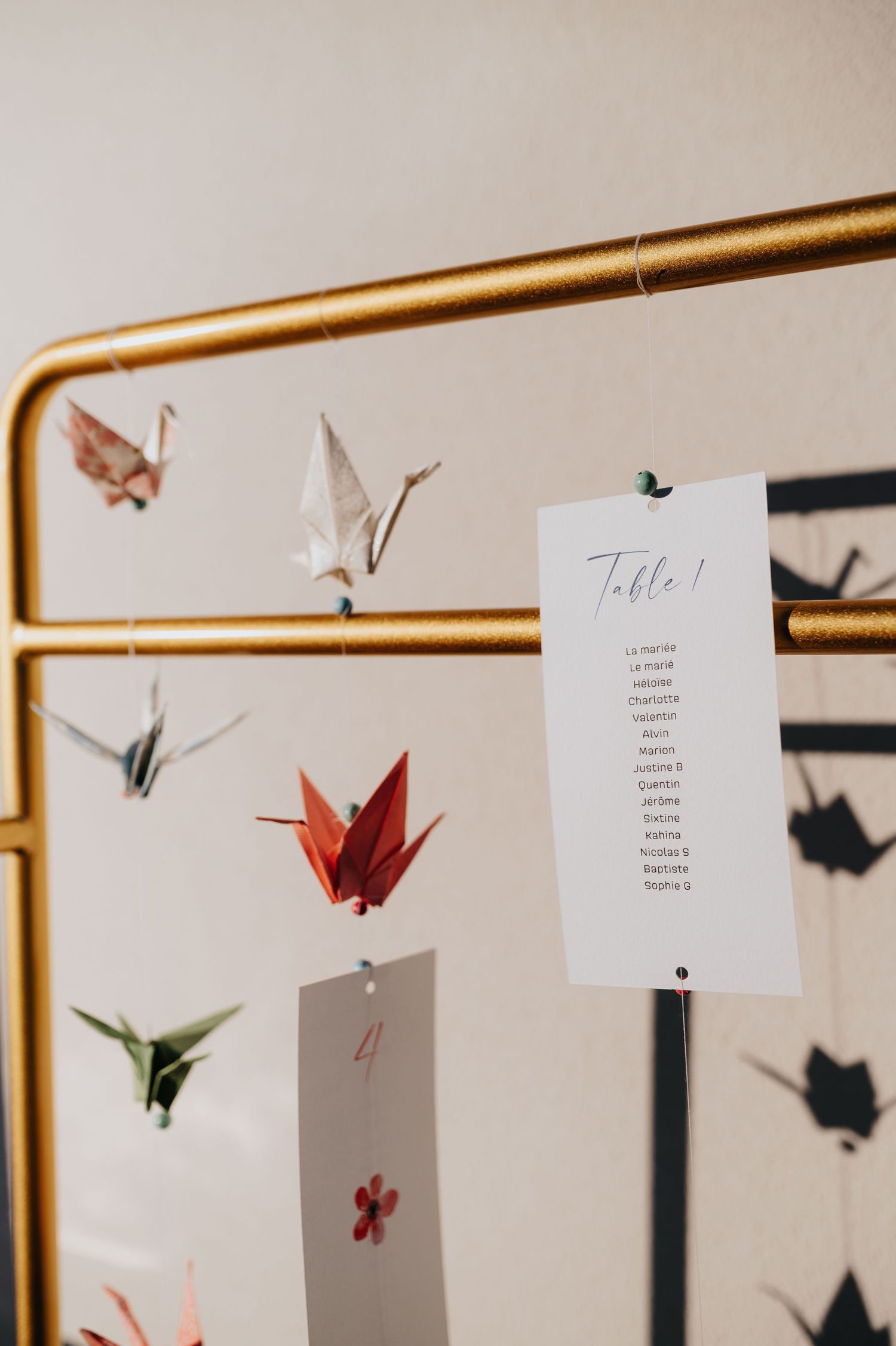 Origami paper cranes and decorative elements hang from a golden rod against a light background.