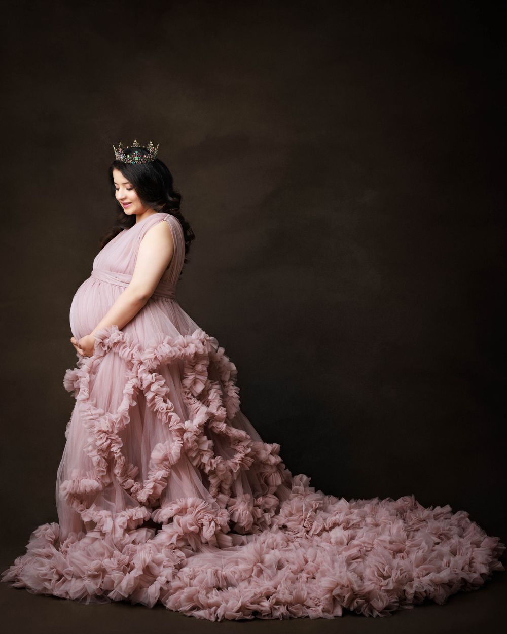 Maternity photo of a pregnant woman in a flowing pink ruffled gown and crown against a dark background.
