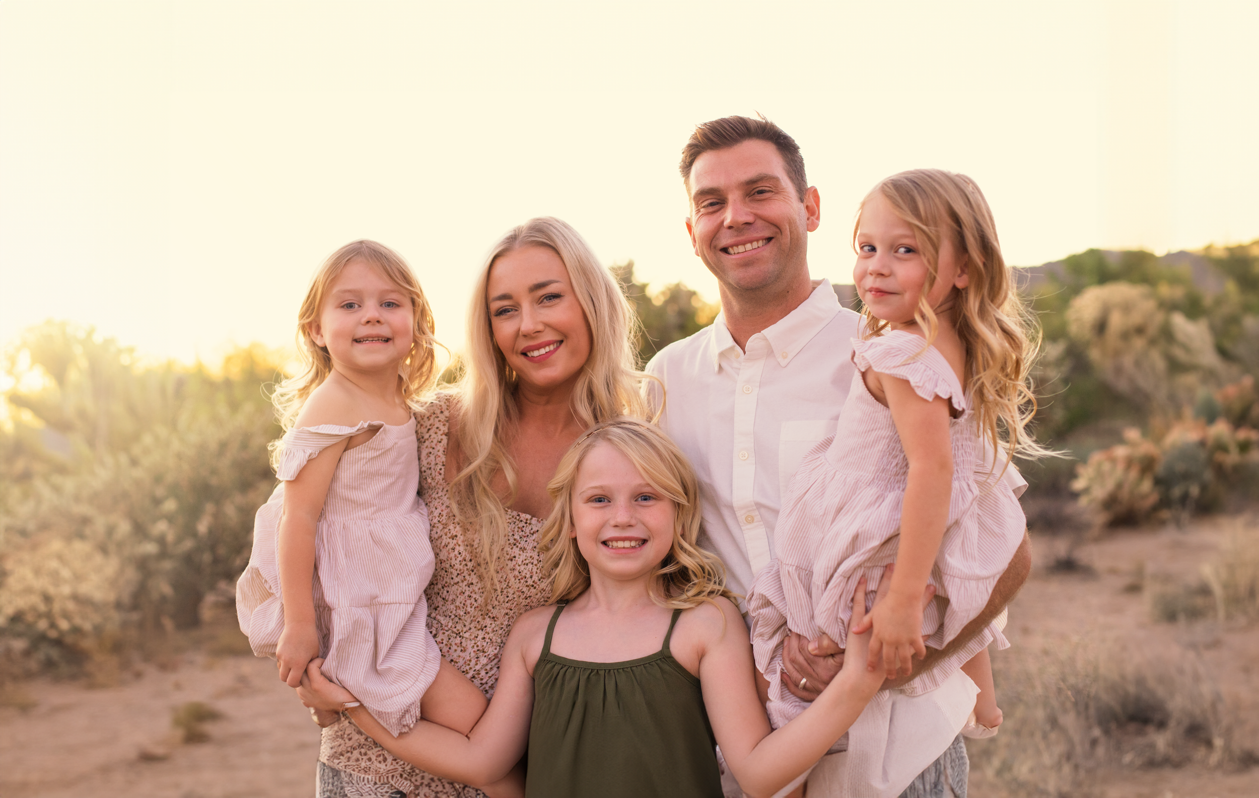 A family wearing coordinated white and neutral outfits poses together in a desert landscape at sunset.