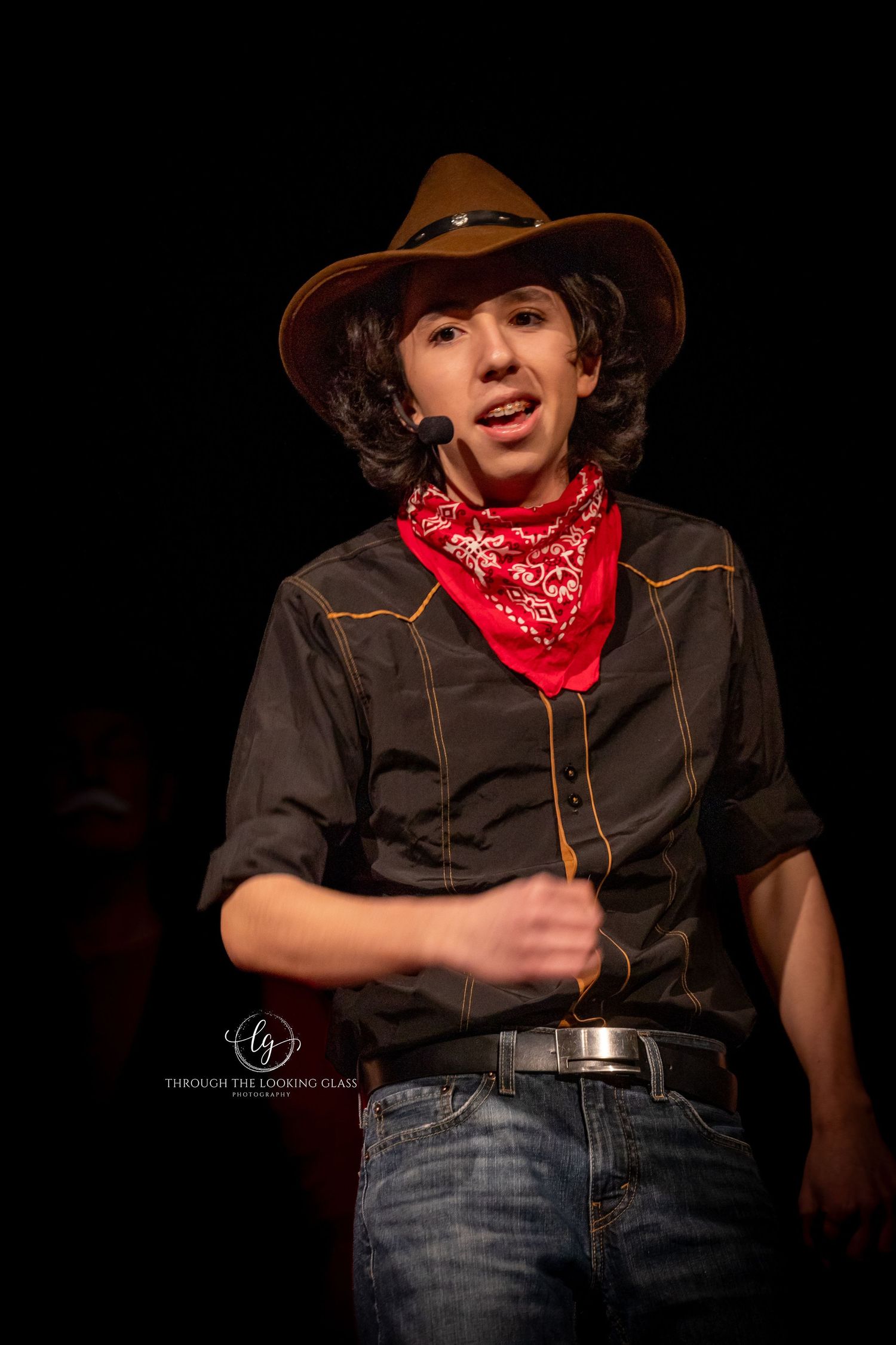 Performer in cowboy hat and bandana delivers lines during Western-themed stage production.