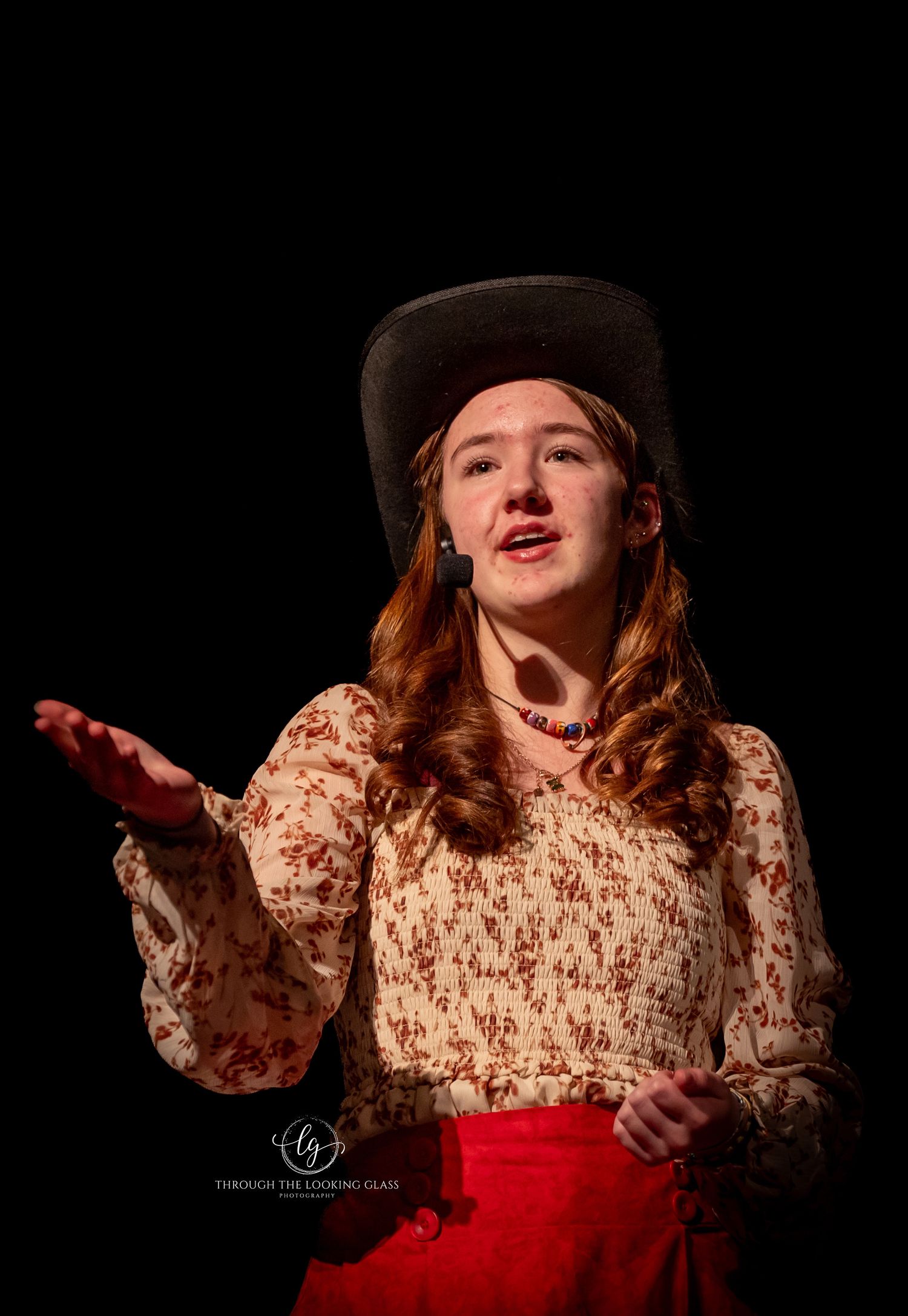 Stage performer in western costume with cowboy hat gestures while singing on dark stage.
