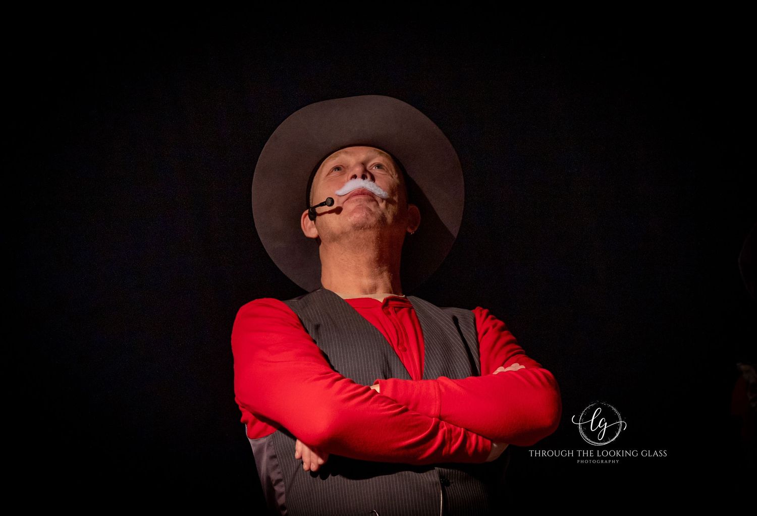 Actor in Western costume with red shirt, vest and cowboy hat performs dramatically against dark background.