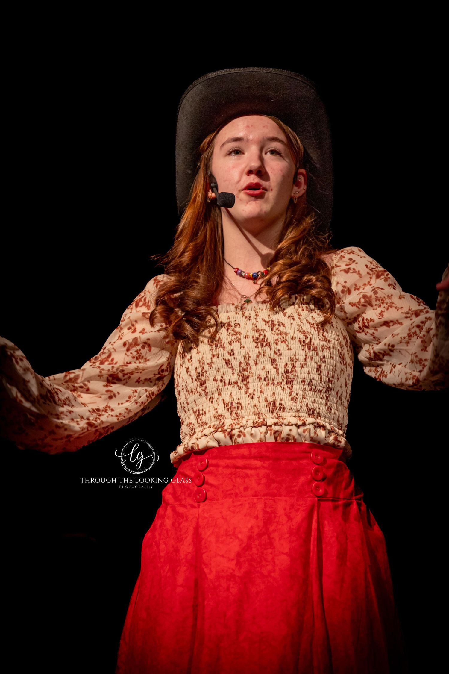 Theatrical performer in period costume with floral top and red skirt gestures expressively on dark stage.