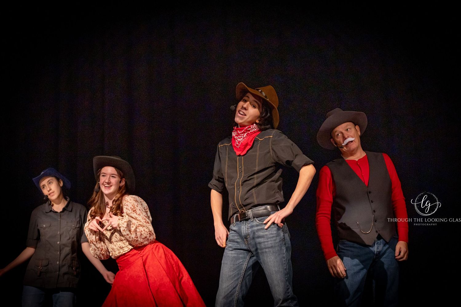 Group of performers in Western-themed costumes pose dramatically on dark stage.