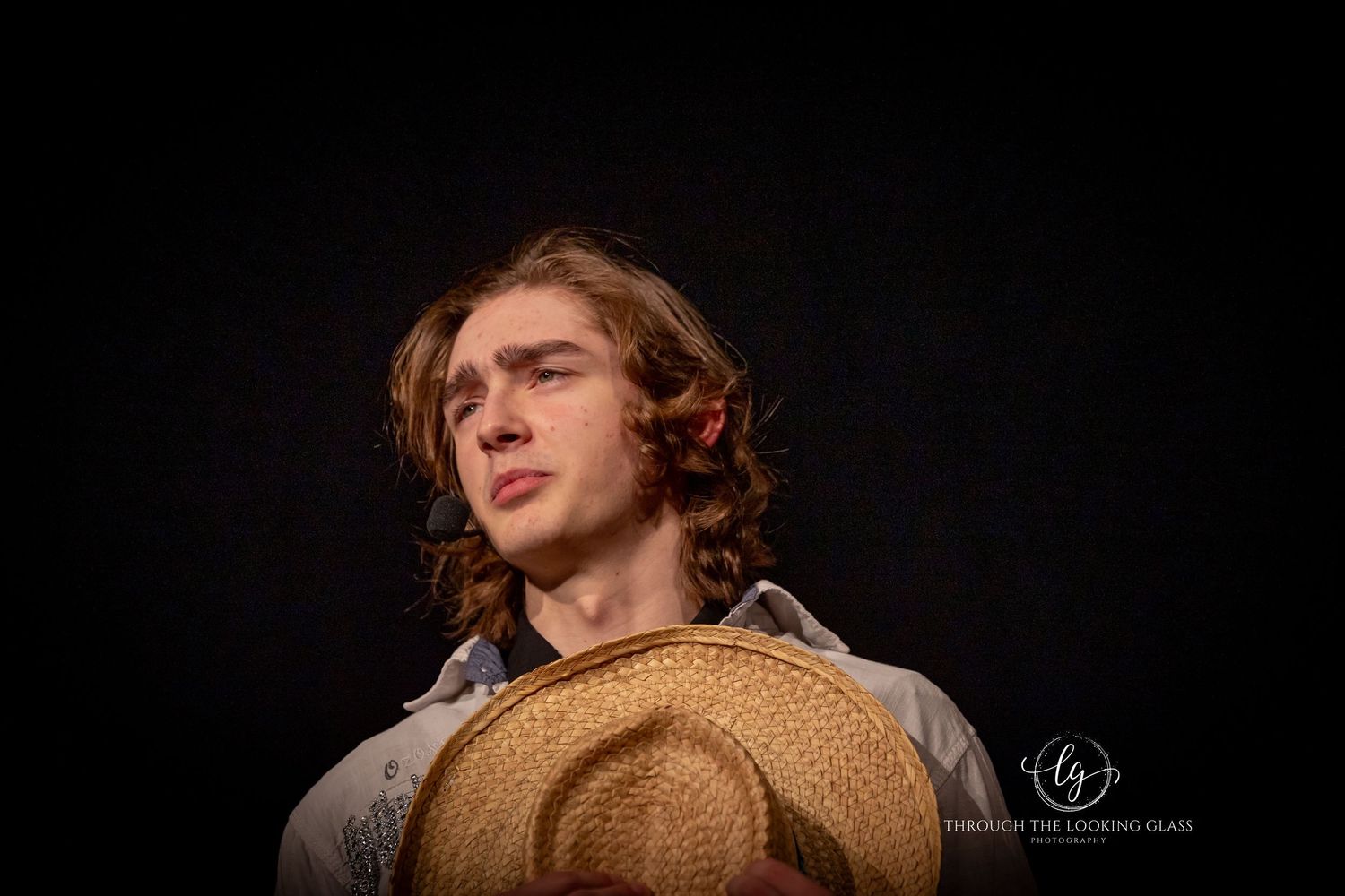 Performer with straw hat poses dramatically against dark background during theatrical production.