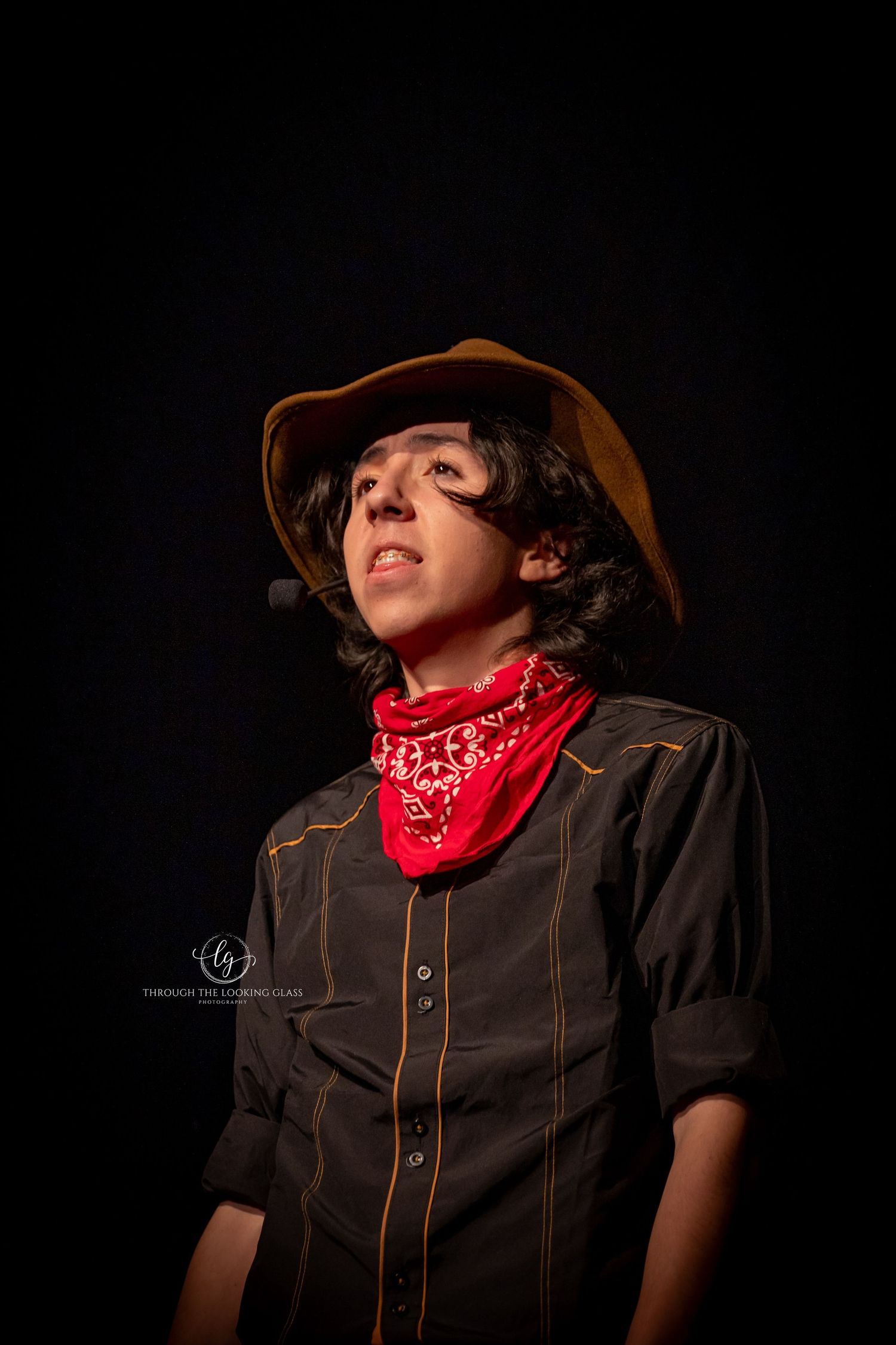 Western-styled performer in dark shirt and red bandana poses dramatically against black backdrop.