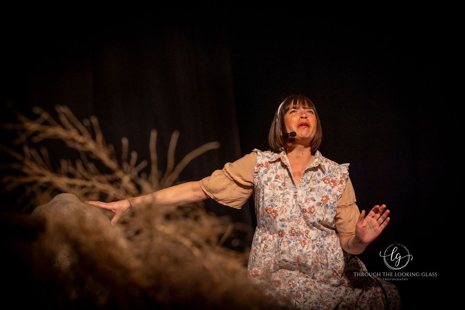 Performer in floral dress gestures dramatically on stage with rustic set pieces.