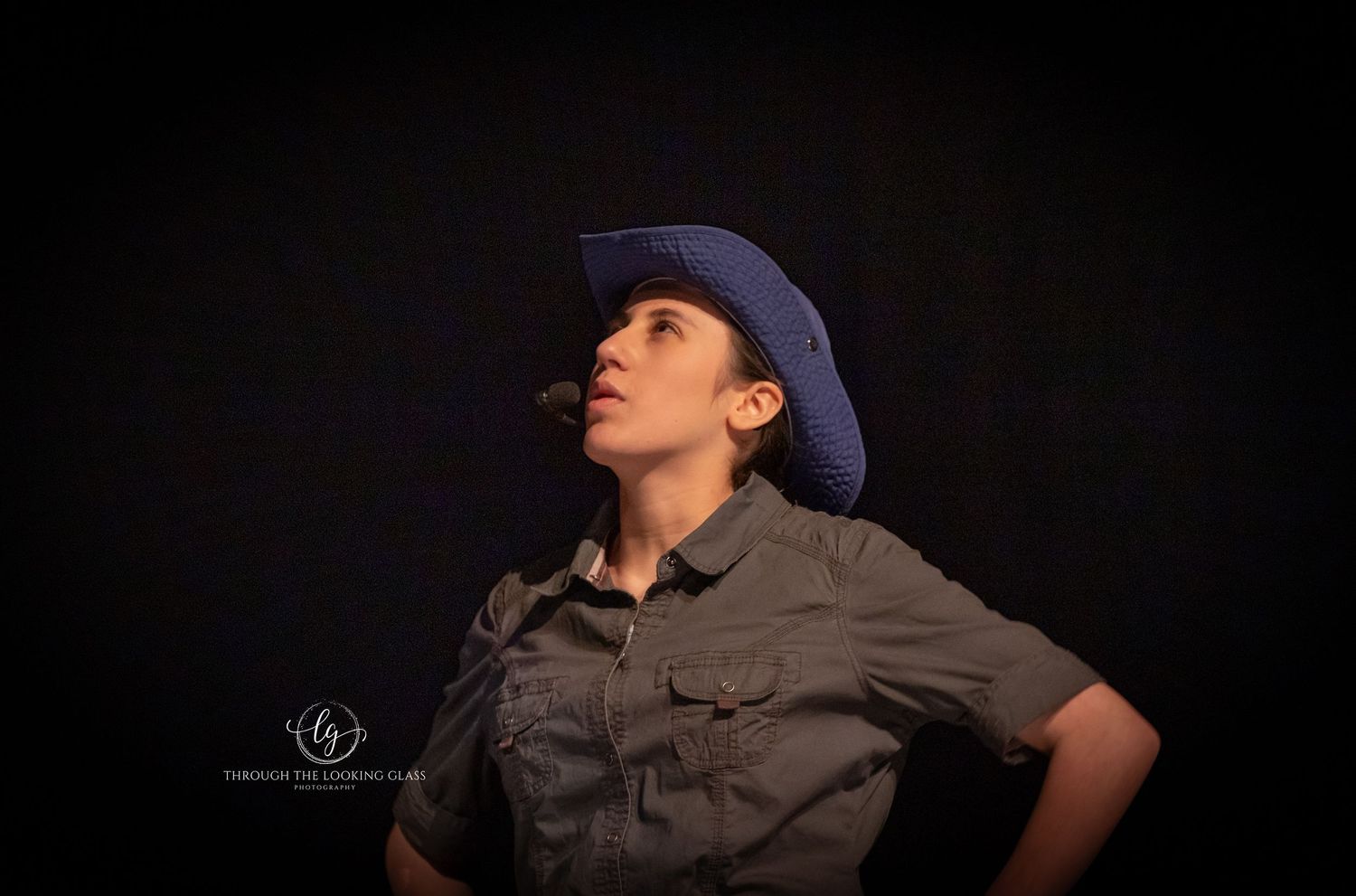 Performer in blue baseball cap and gray shirt poses dramatically against dark background.