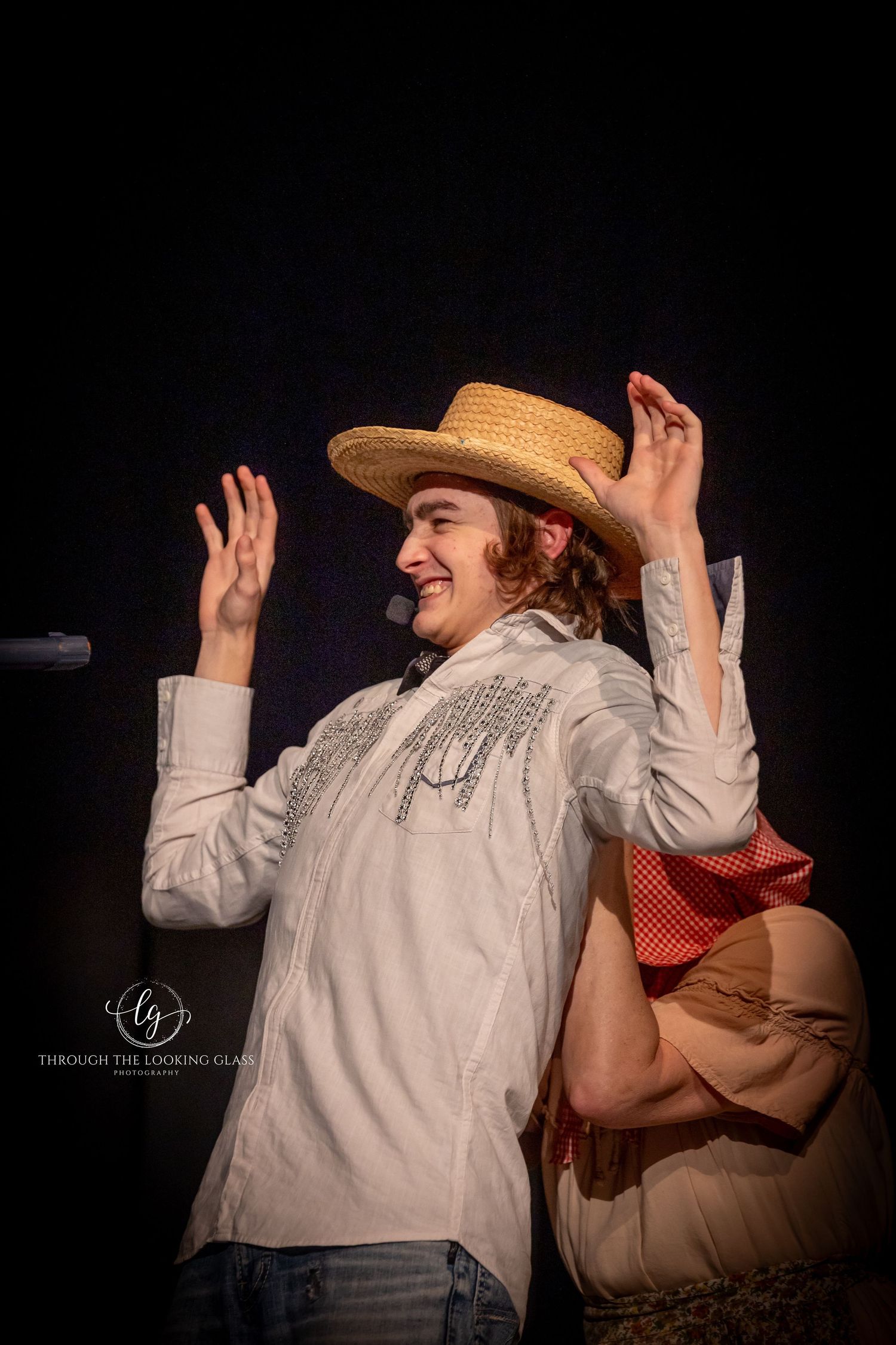 Performer in straw hat and white shirt raises hands energetically during theatrical scene.