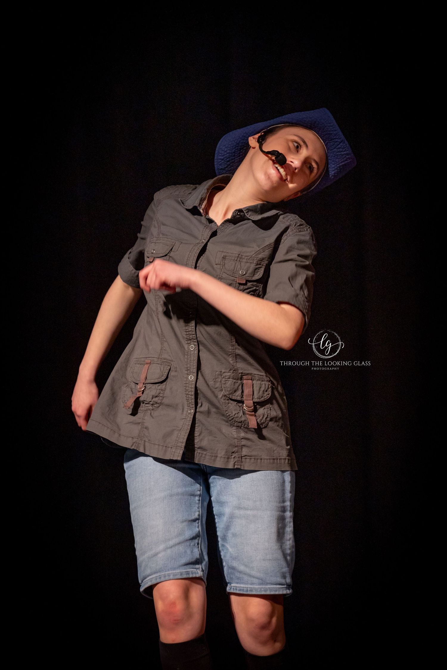 Actor in blue cap and utility jacket performs expressive pose against dark backdrop.
