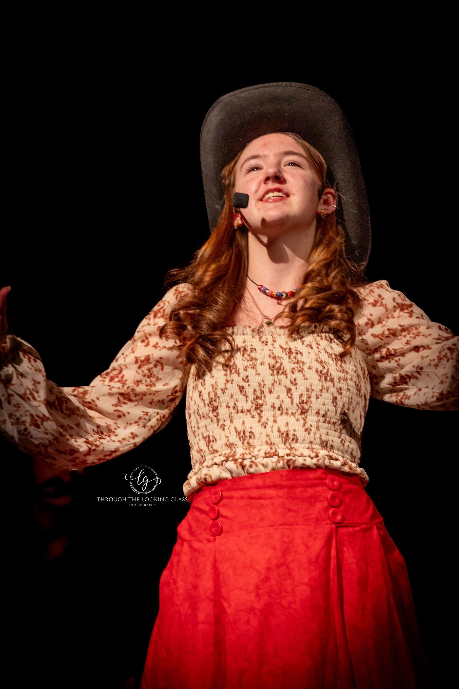 Theatrical performer in western costume with floral dress and cowboy hat gestures dramatically.