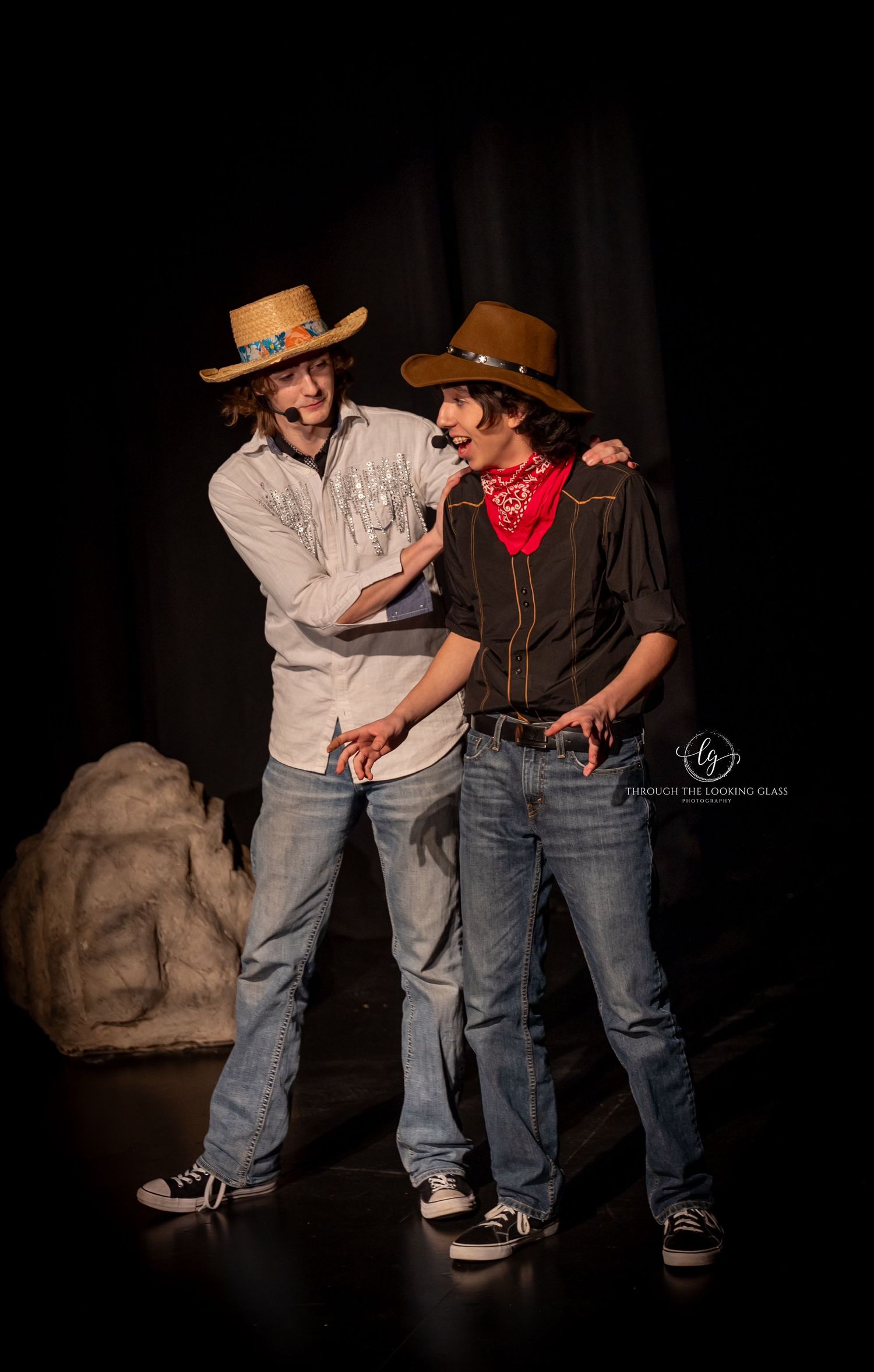Two western-styled performers interact dramatically on stage with rustic backdrop.