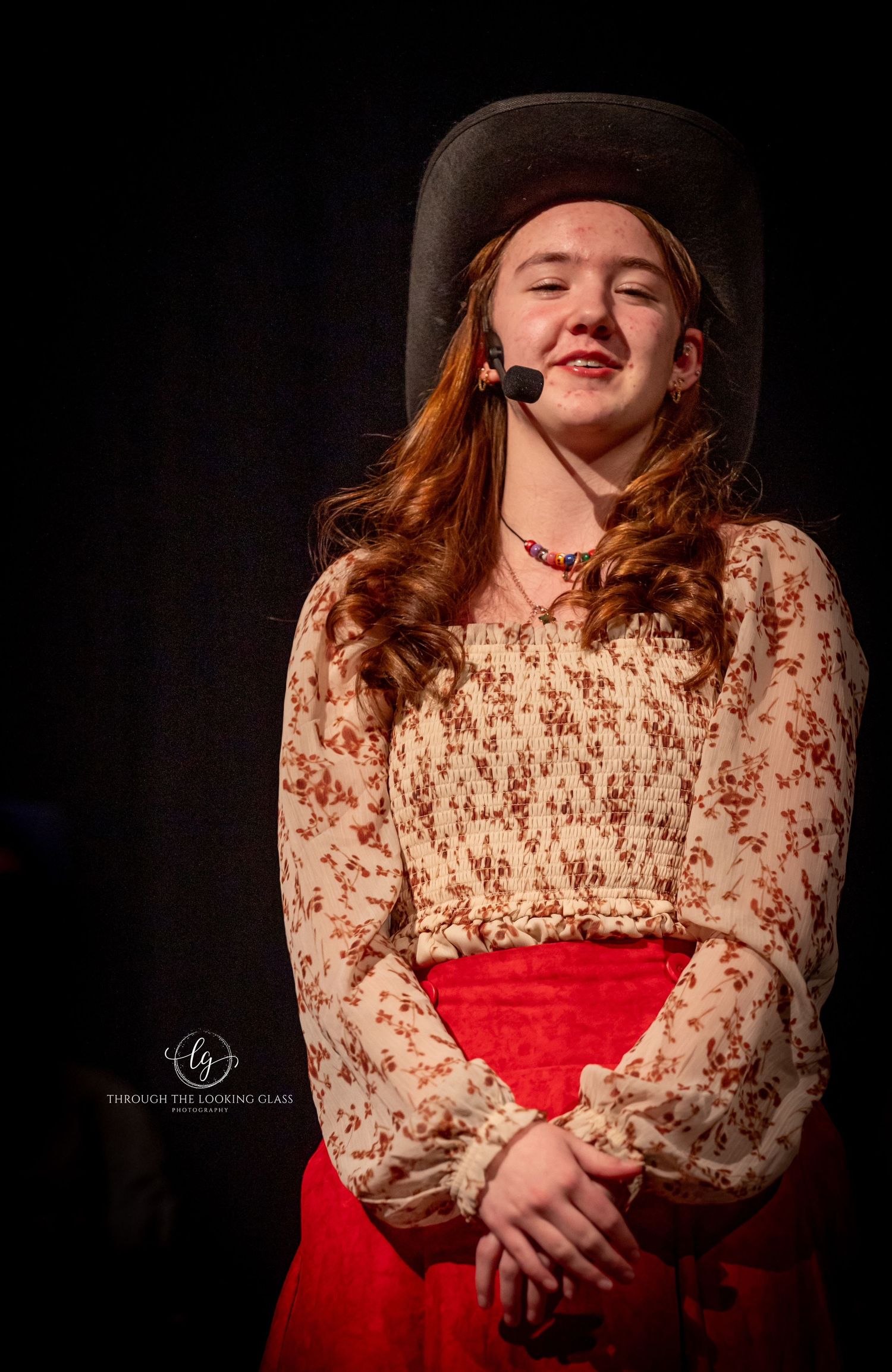 Performer in western attire including brown hat and floral dress with red skirt performs on dark stage.
