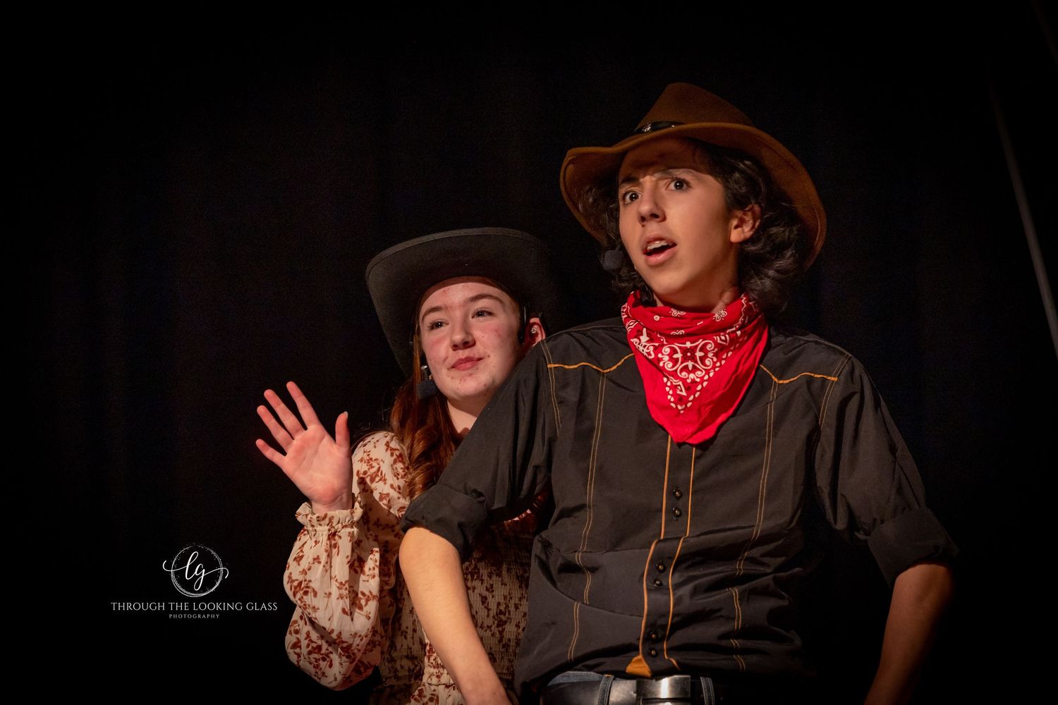 Two performers in western costumes during theatrical production on dark stage.