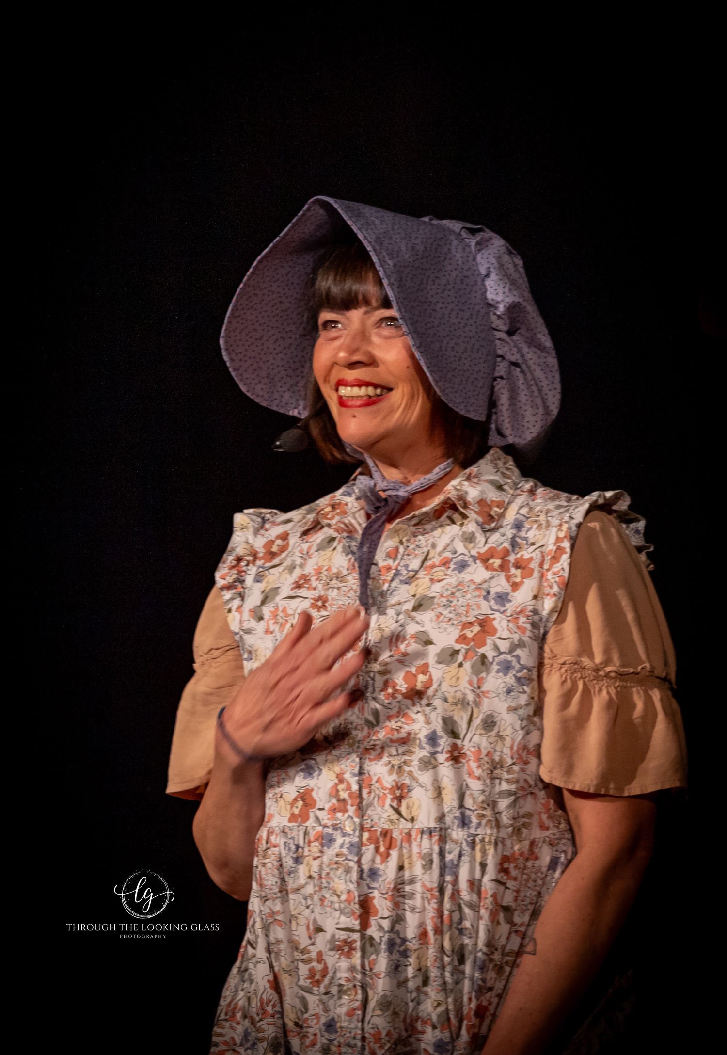Performer in period bonnet and floral dress laughs joyfully during theatrical scene.