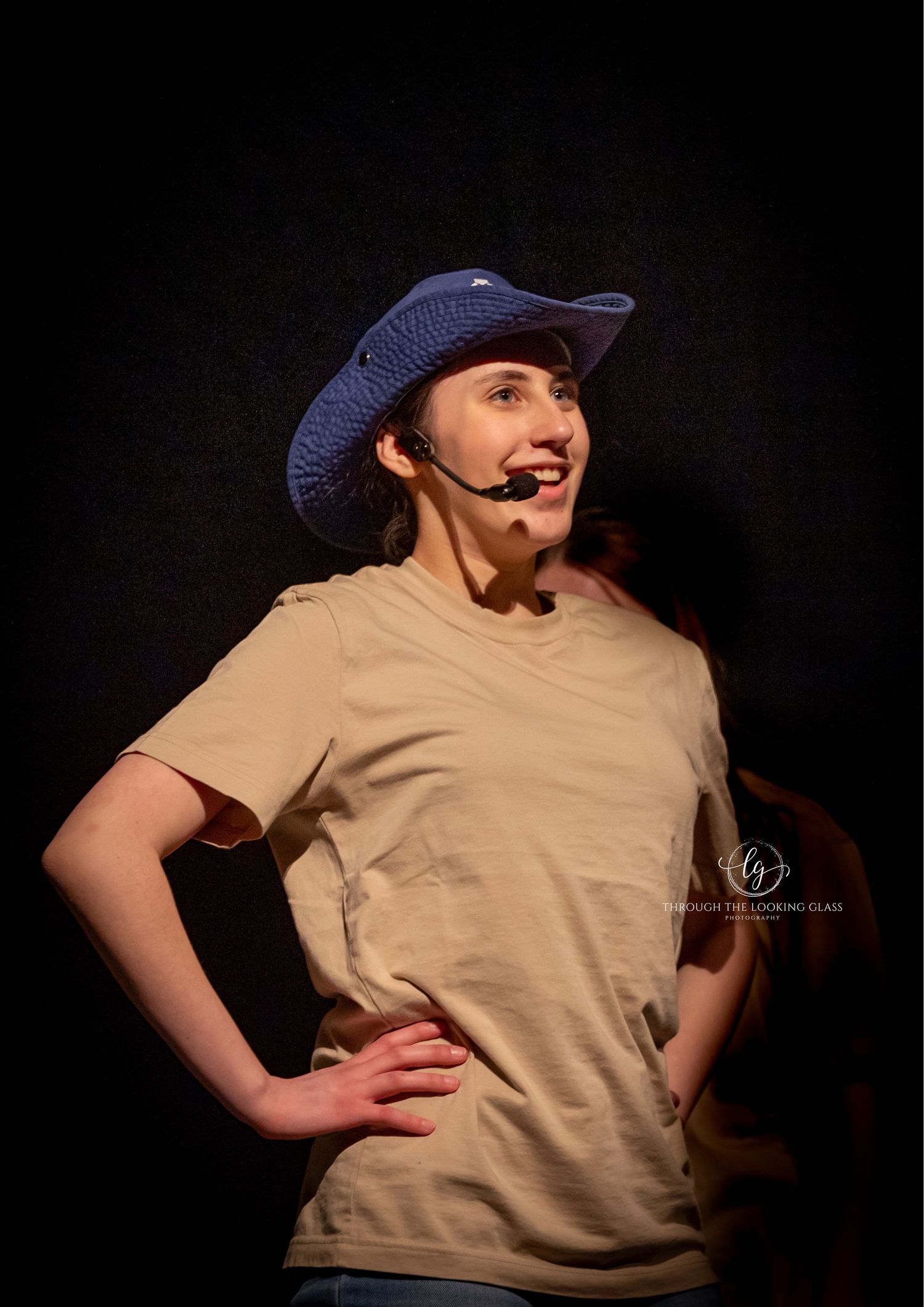 Performer wearing blue cowboy hat and tan shirt on dark stage with microphone.
