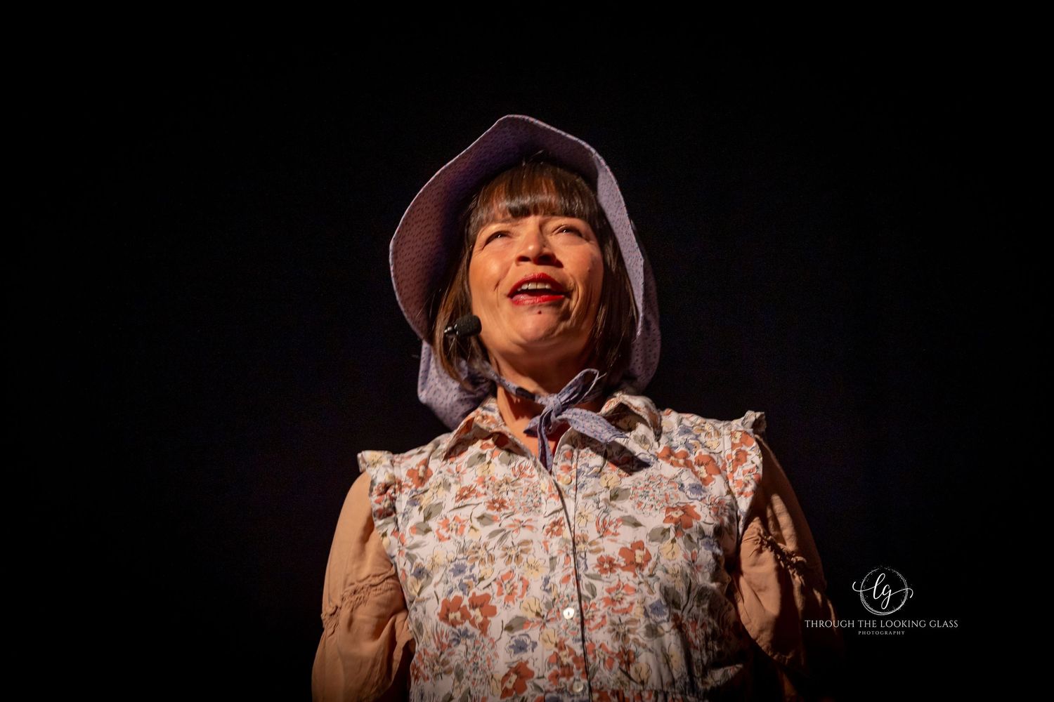 Theatrical performer in brown hat and floral print clothing expressing joy on dark stage.