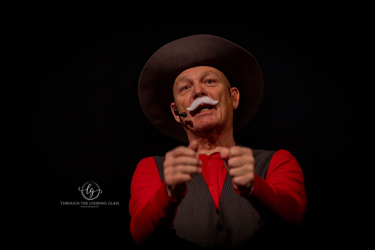 Performer in red shirt and cowboy hat gestures expressively during theatrical scene.