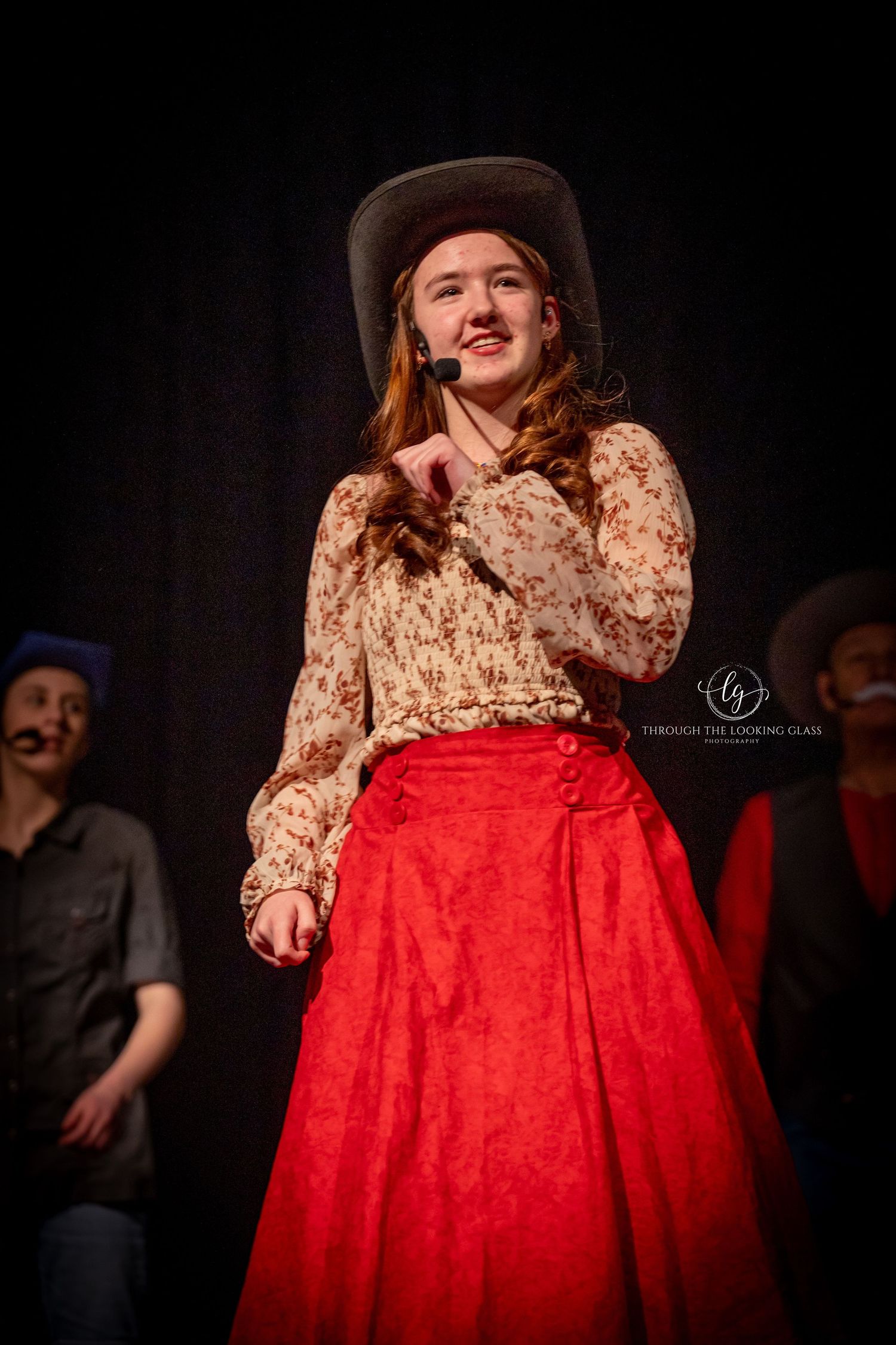 Theater performer in floral top and red skirt with cowboy hat on stage.