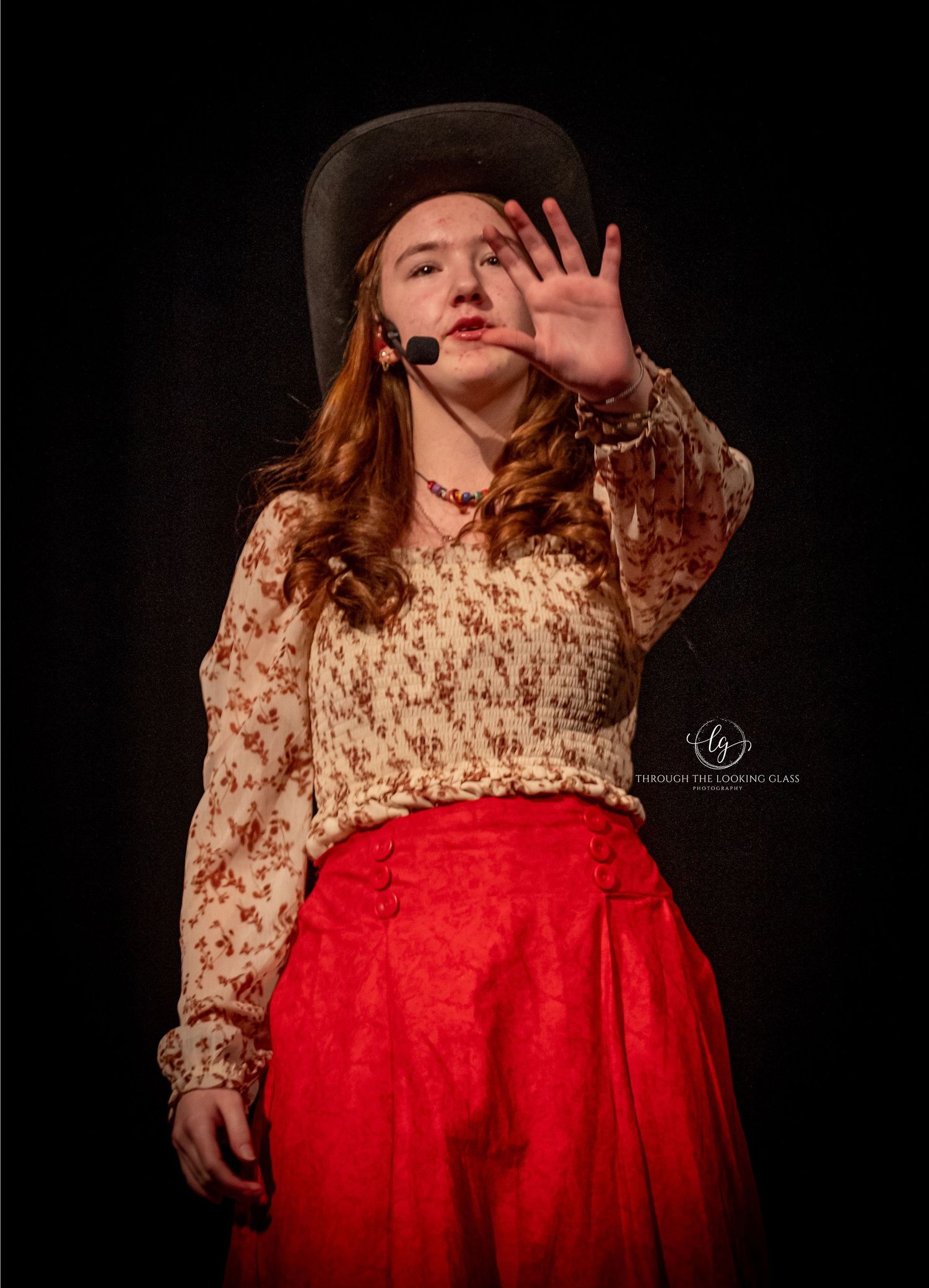 Performer in vintage floral blouse and red skirt gestures dramatically during theatrical show with dark background.