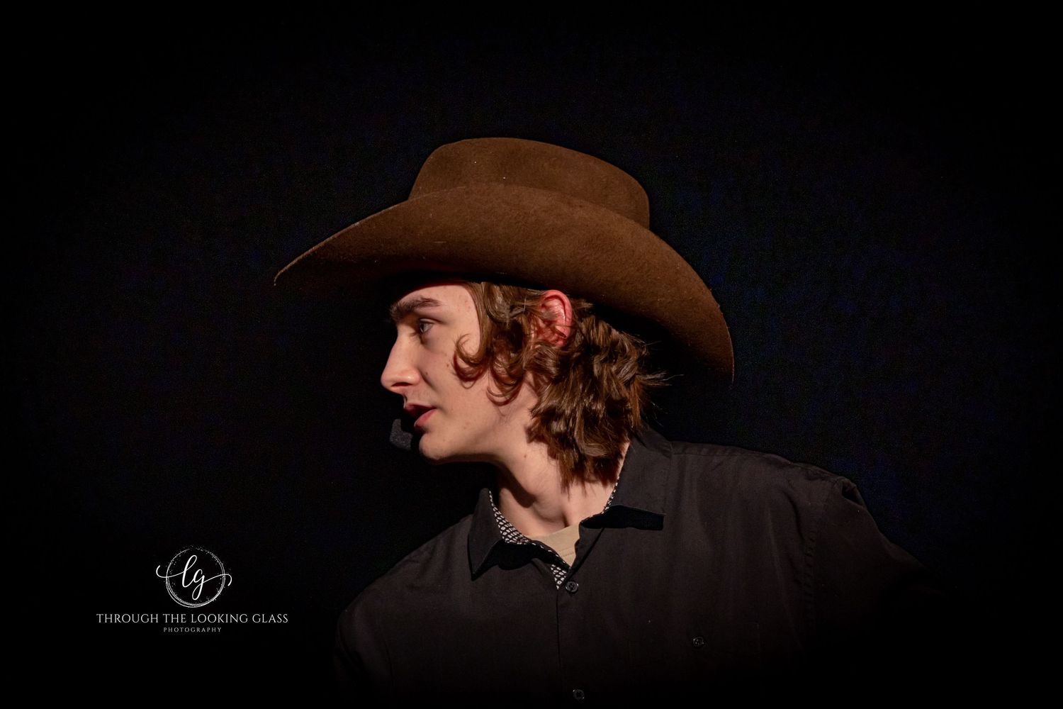 Side profile portrait of western performer in brown cowboy hat against dark backdrop.