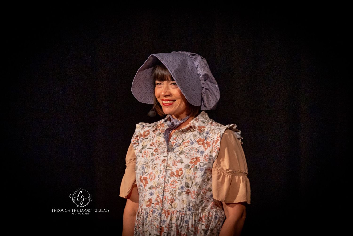 A performer wearing a floral dress and bonnet smiles during a theatrical production.