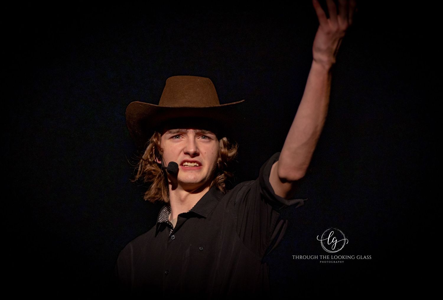 Dramatic stage performer in cowboy hat raises hand against dark background during theatrical performance.