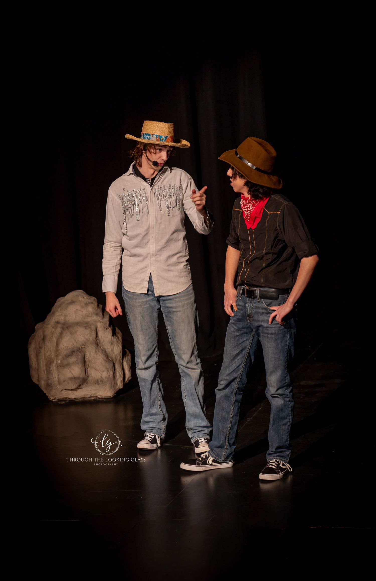 Two performers in western wear and cowboy hats interact on dark stage with prop rock.