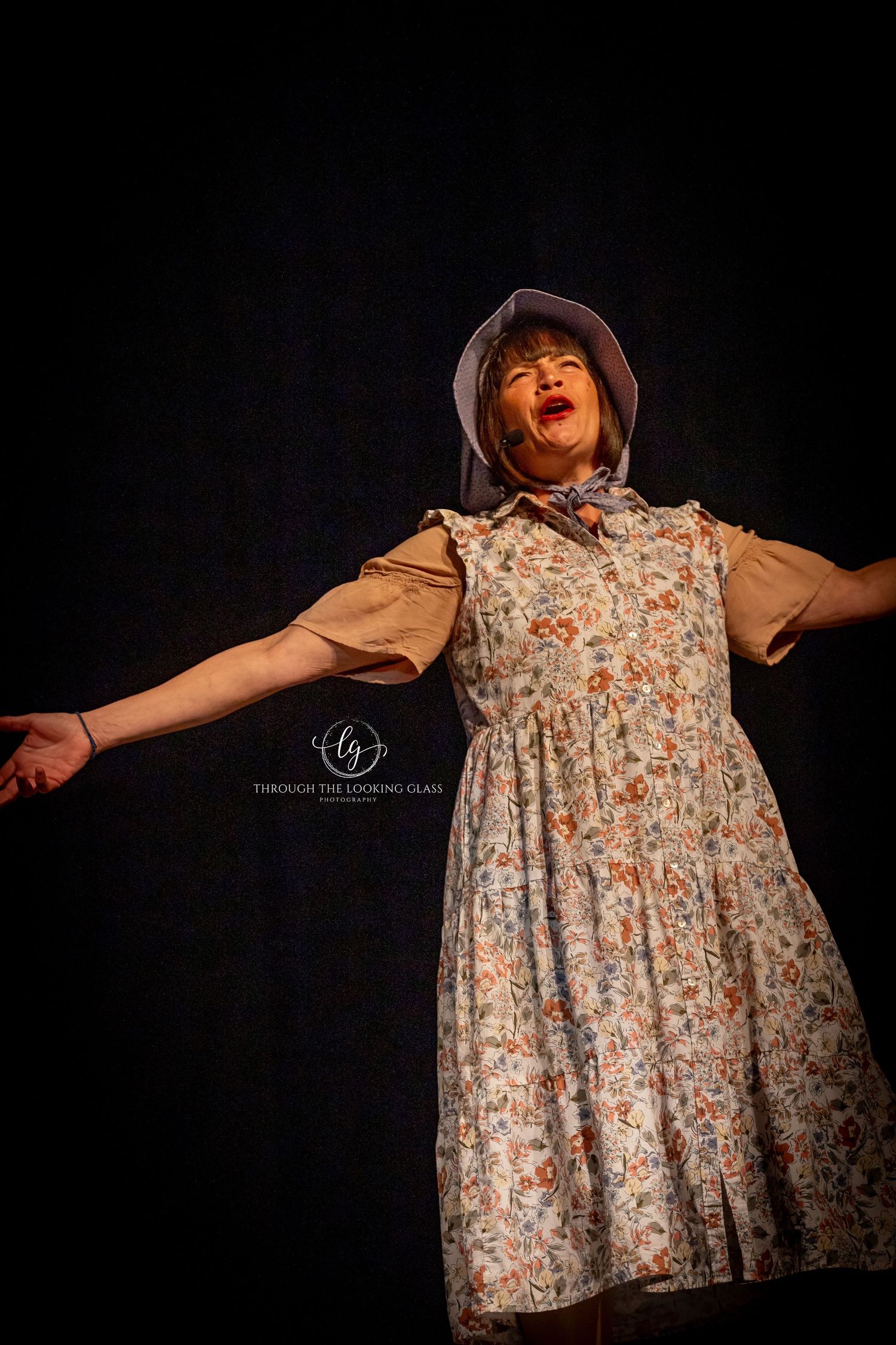 A performer in a floral dress and bonnet spreads their arms wide during an emotional performance.