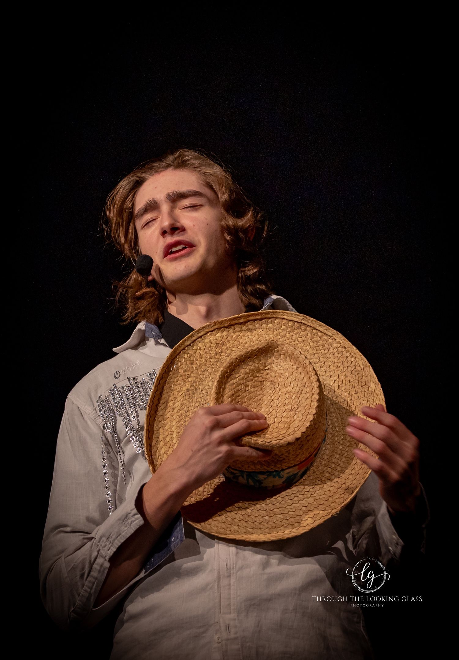Theatrical performer holding straw hat while singing in dramatic lighting.