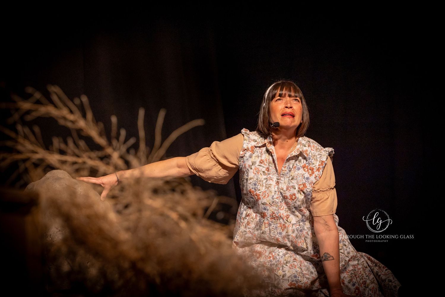 Theater actor in floral dress sitting on rustic set with dramatic lighting.