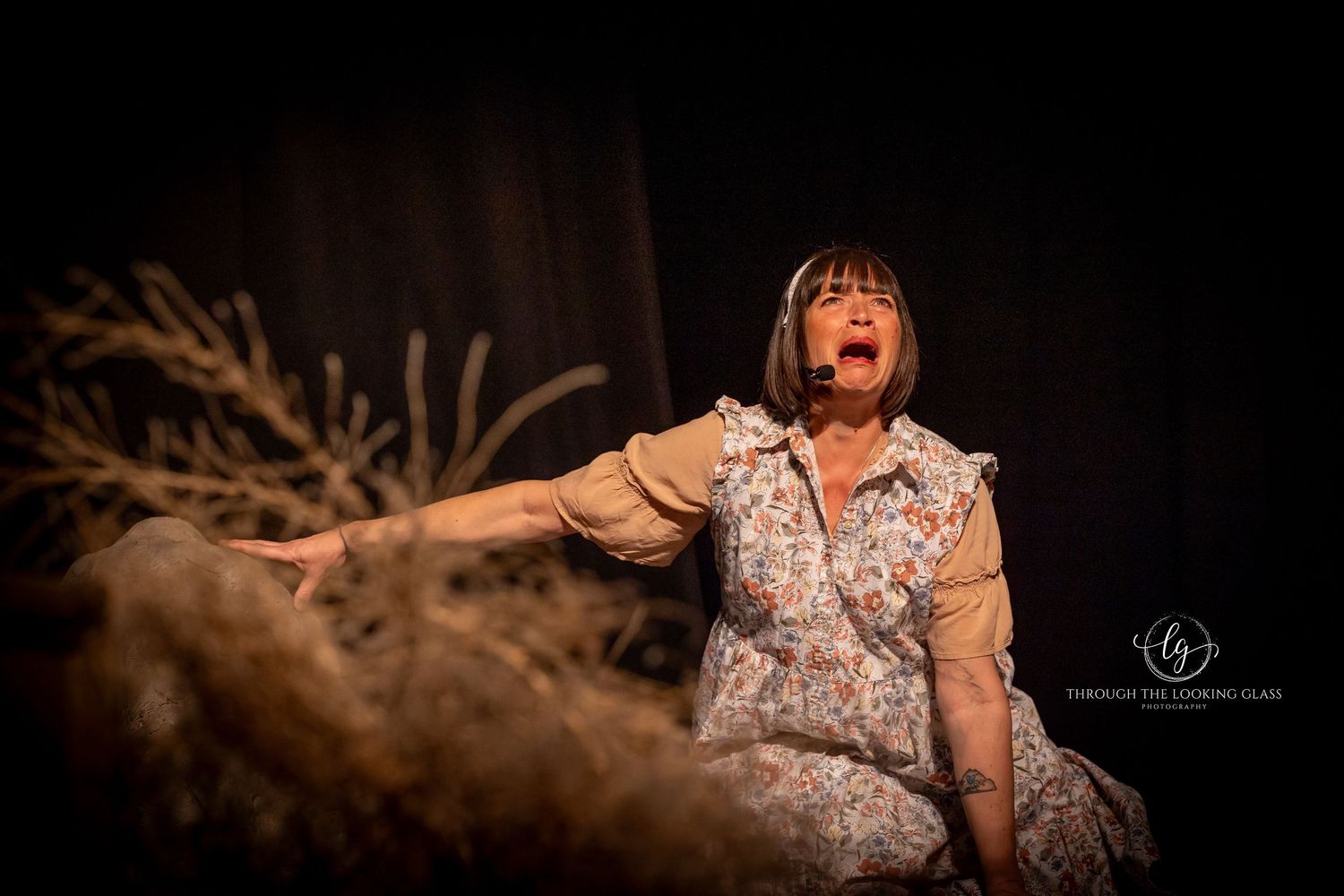 A theatrical performer in a floral dress dramatically gestures during a stage performance against a dark background.