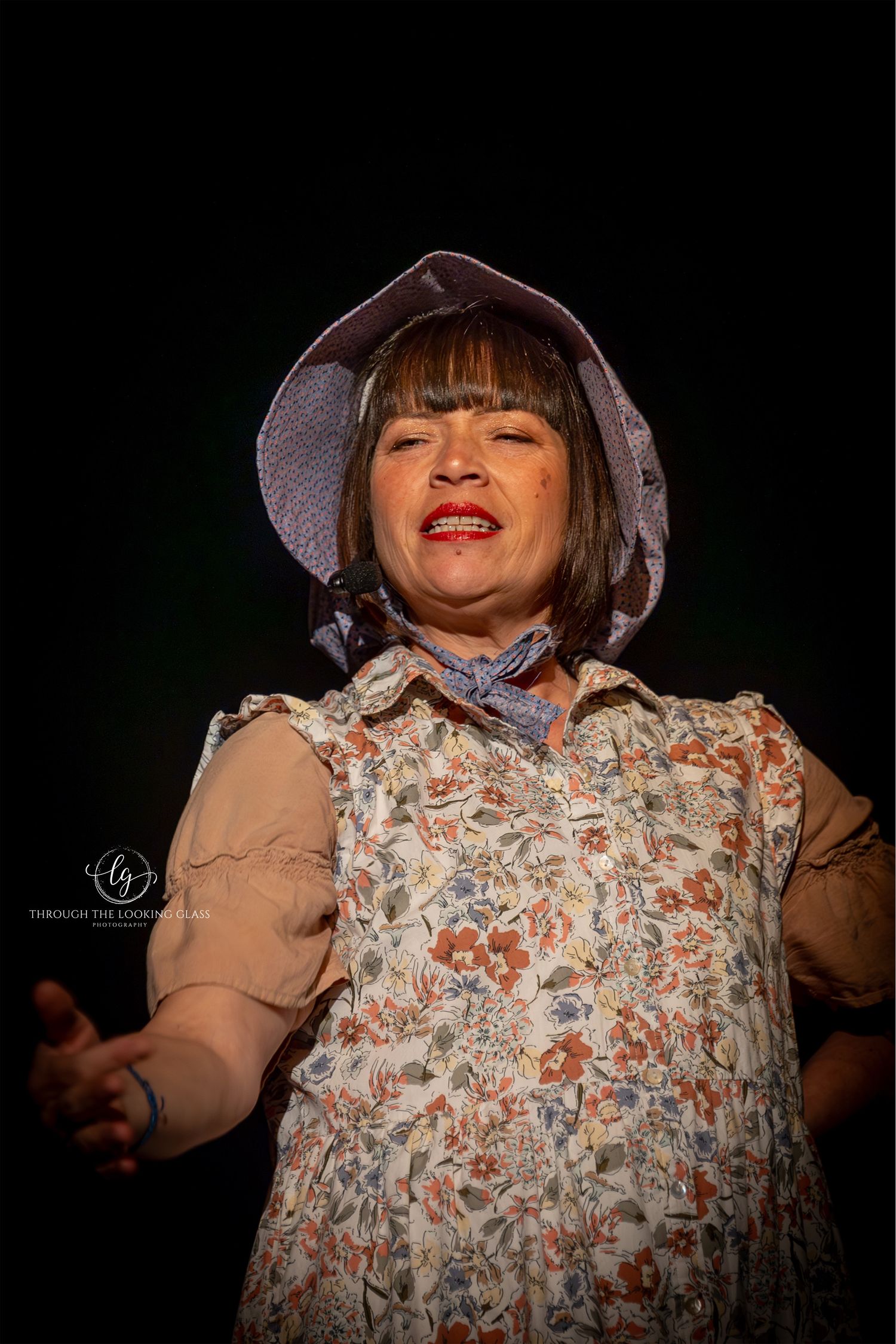 A performer wearing a floral dress and bonnet sings expressively on a dark stage.
