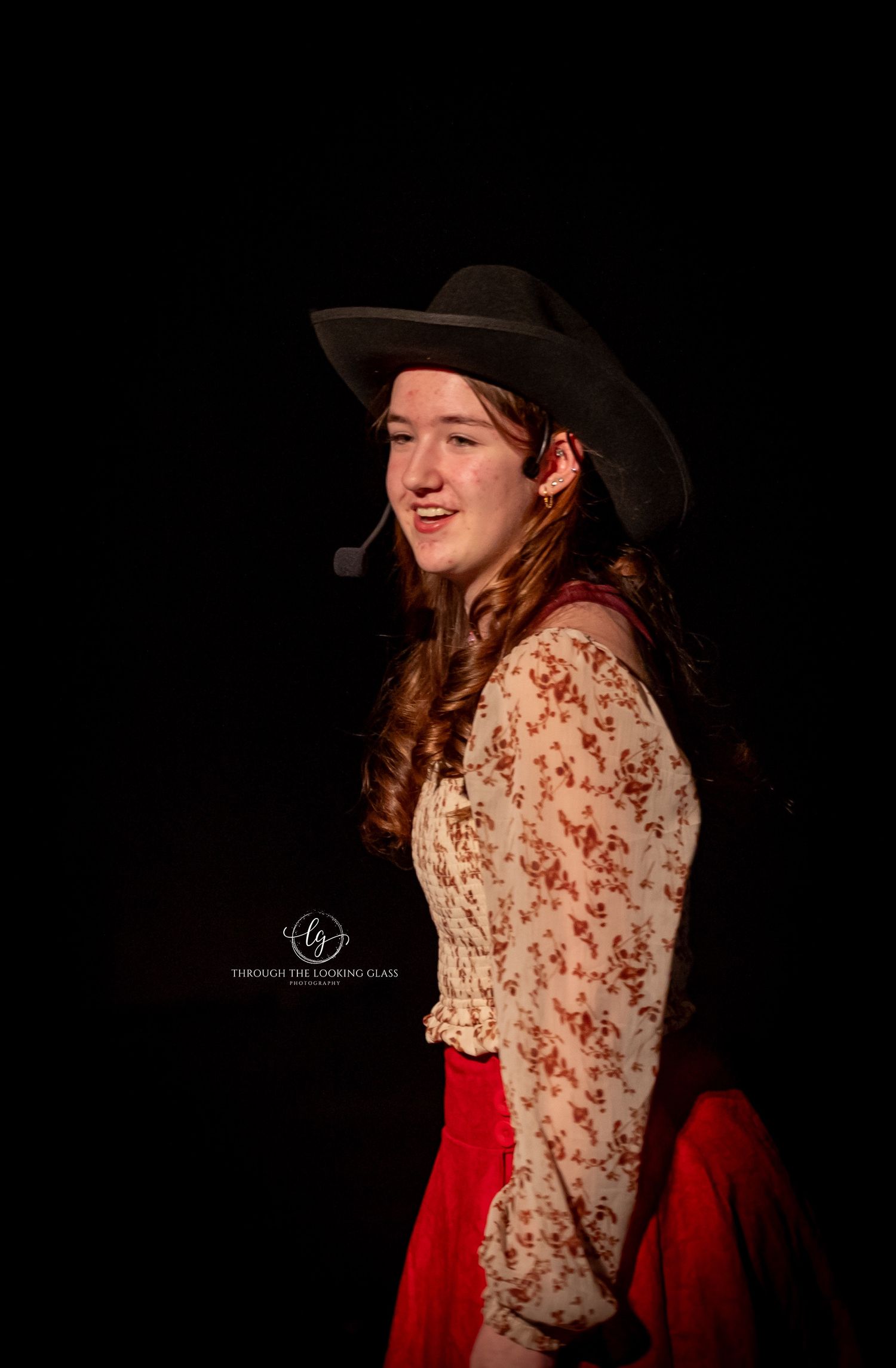 A performer in western attire including cowboy hat and red skirt smiles during a stage production.