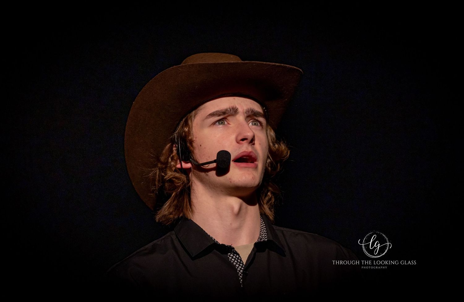 A dramatic stage performer with a cowboy hat and headset microphone performs against a dark backdrop.