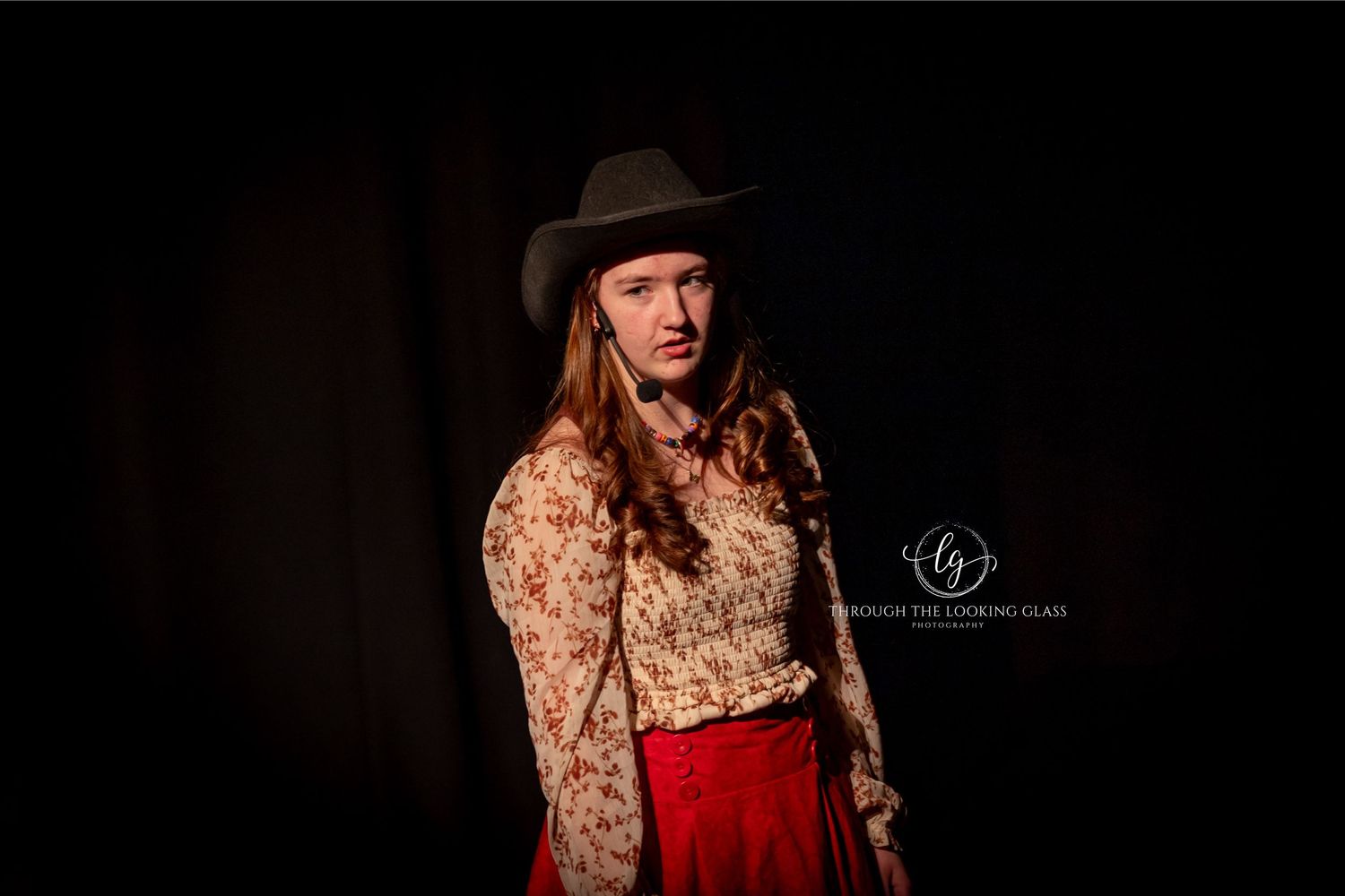 A performer in western costume with long hair poses dramatically against a dark background.