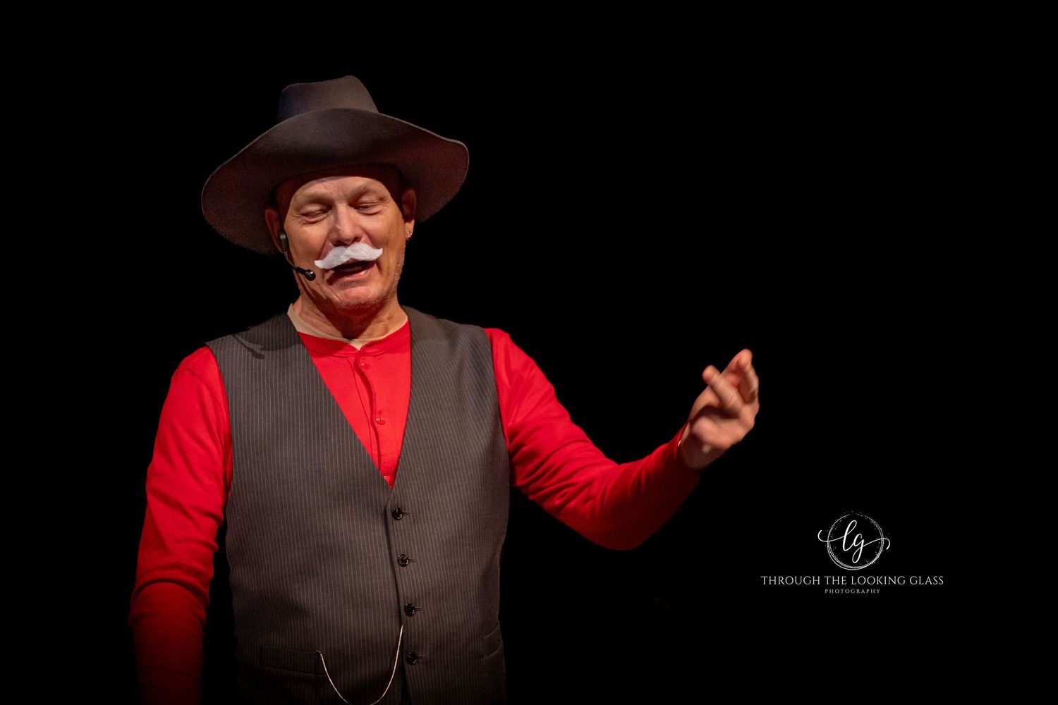 Western-styled performer in a brown hat and red shirt gestures dramatically against a dark background.