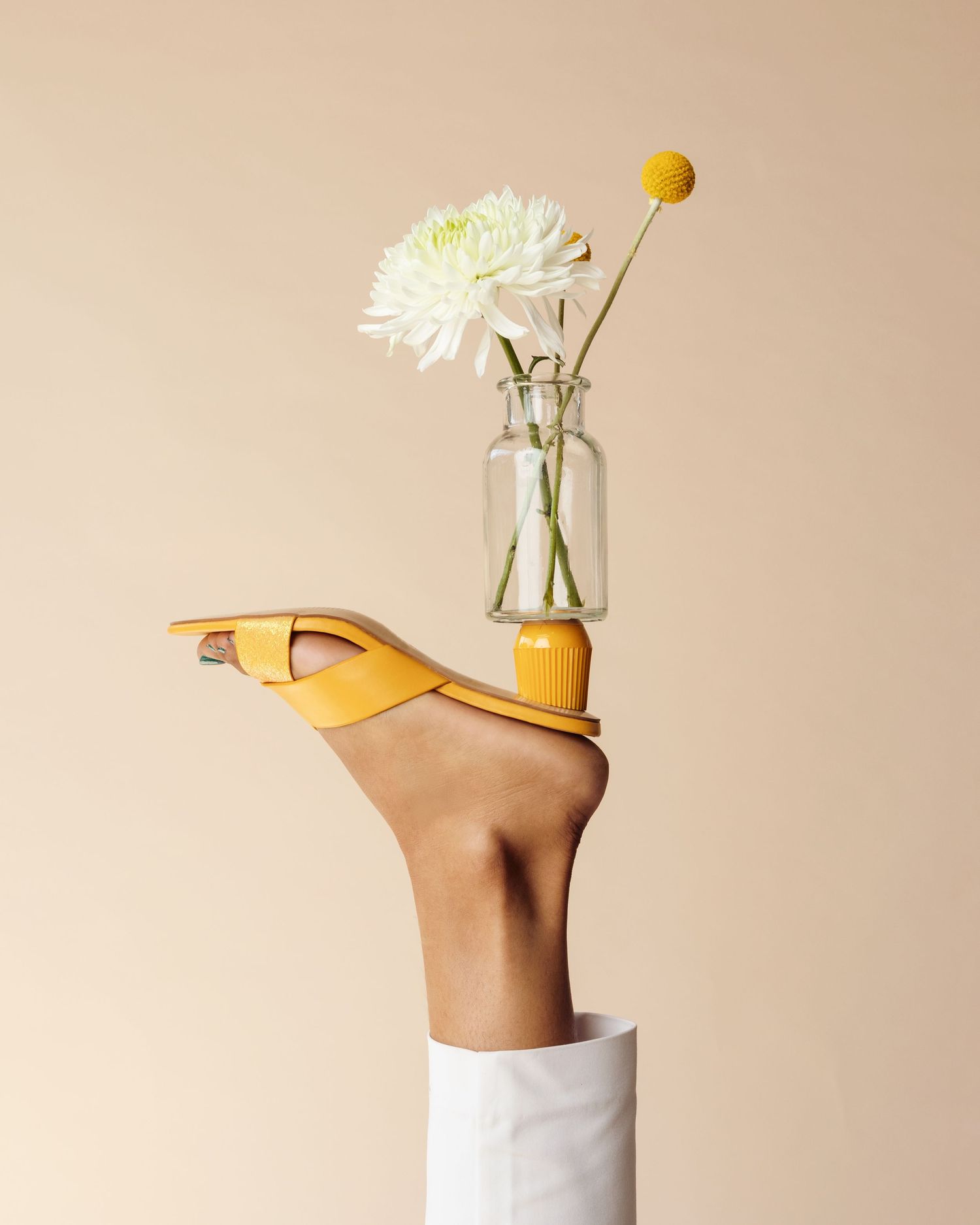 White dahlia and yellow craspedia flowers in glass vase balanced on tan heel shoe against beige background.