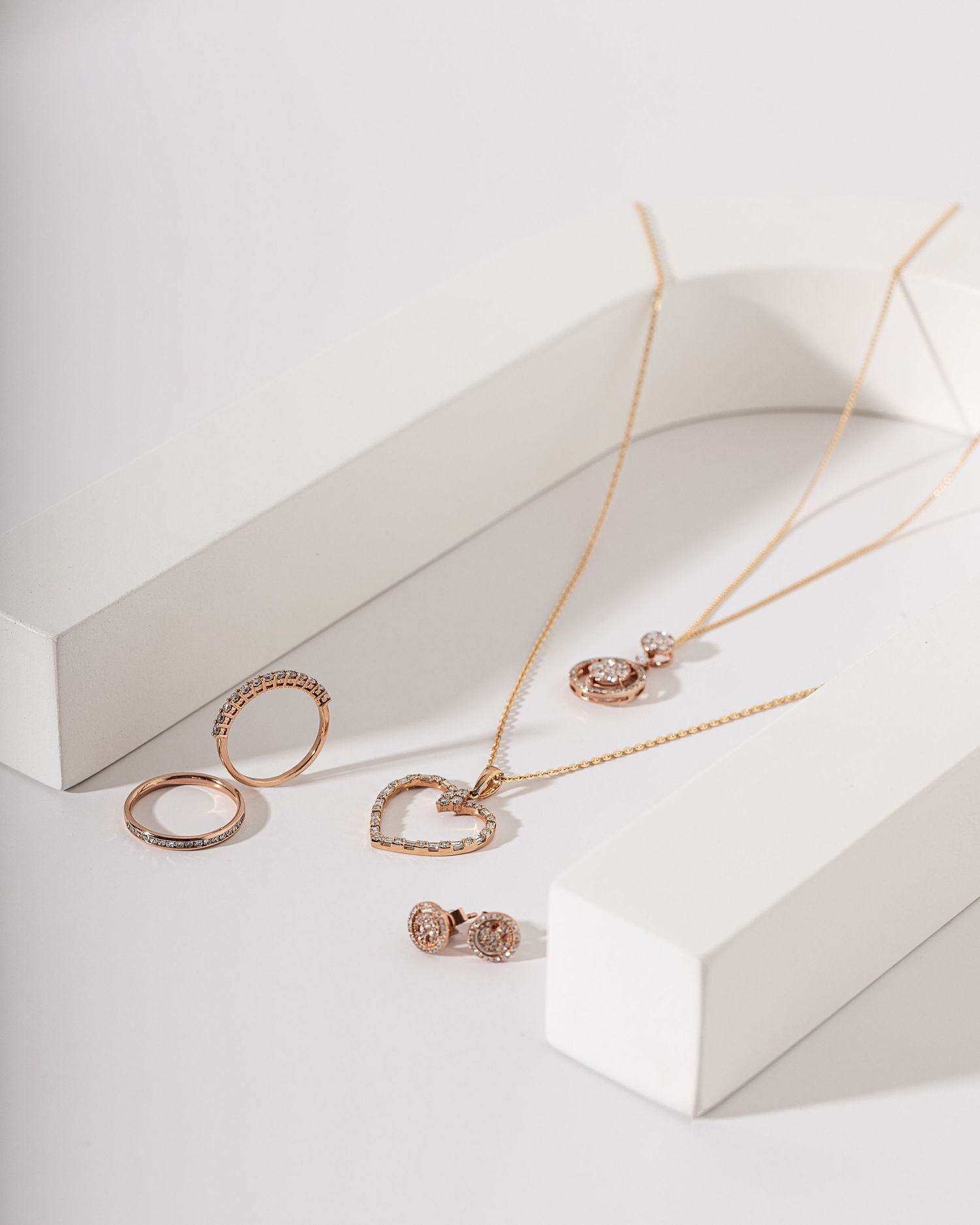 Rose gold jewelry collection featuring rings and necklaces displayed on white geometric platforms.
