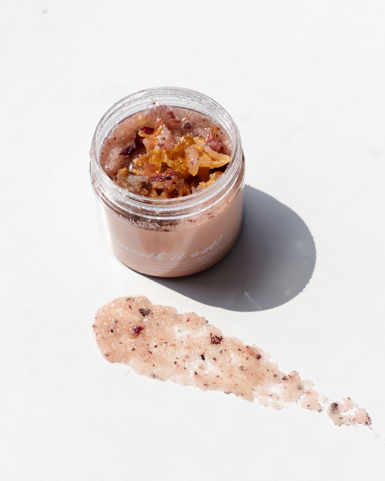 Brown body scrub in glass jar with product swatch smear on white surface.