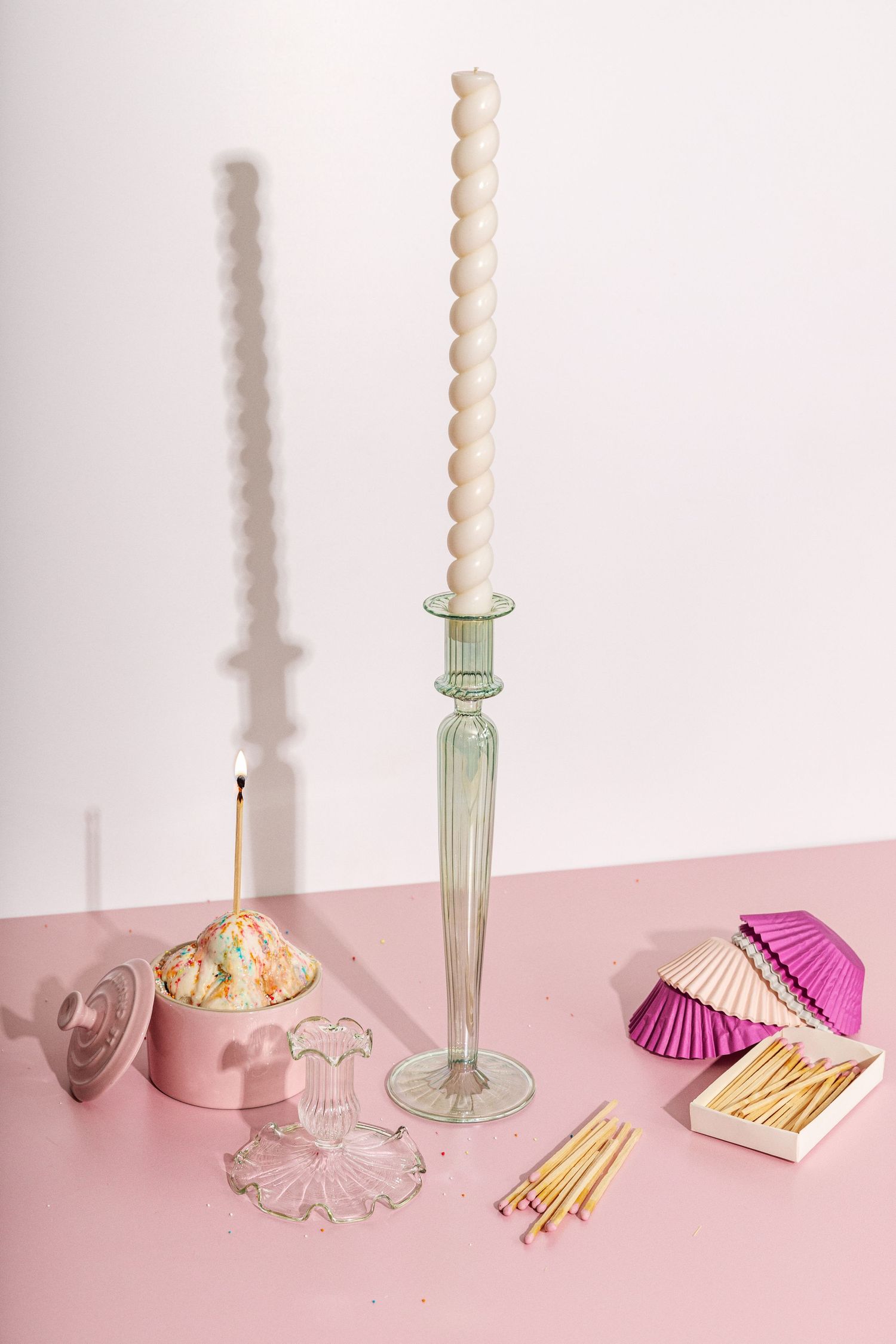Elegant glass candlestick with spiral candle and pink party accessories on pink surface.