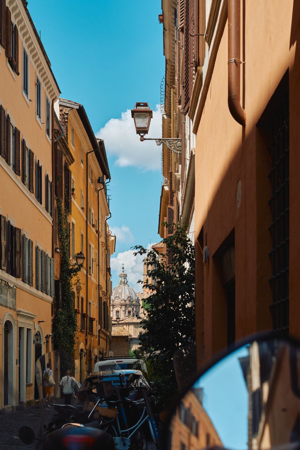 Narrow Italian street with warm-colored buildings and a church dome visible in the distance against bright blue sky.