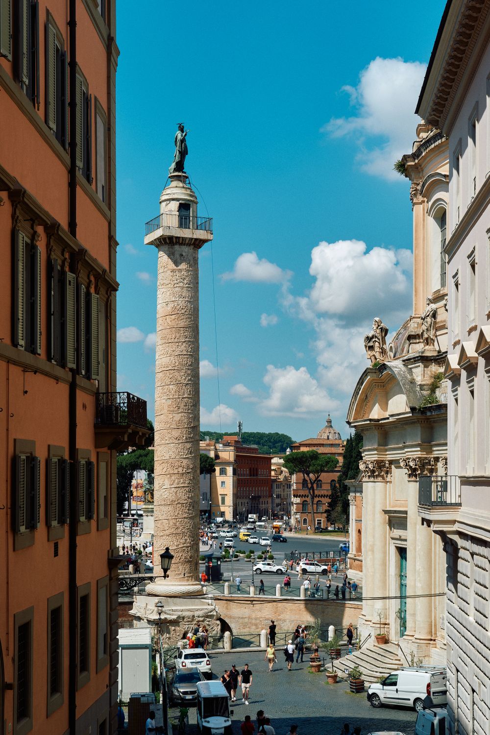 Tall Roman column stands prominently in a bustling Italian city square with historic buildings and parked cars.