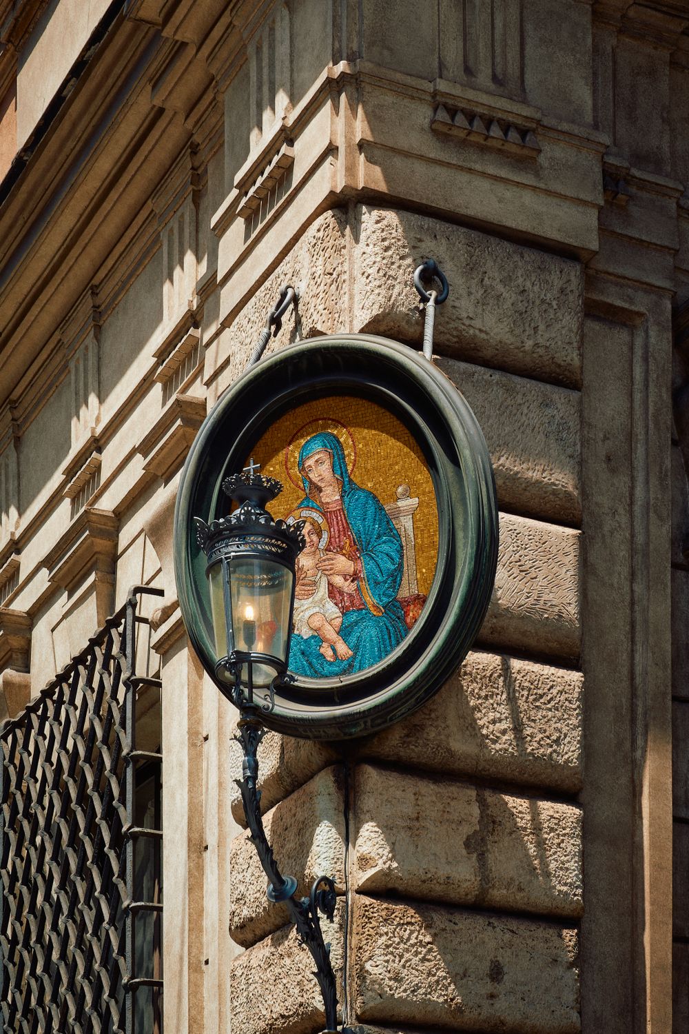 Ornate mosaic of Madonna and child within a circular frame mounted on a historic stone building wall with decorative lamp.