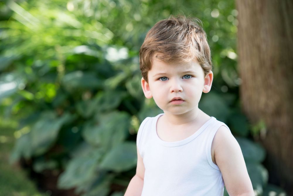 Young child in white tank top standing outdoors against blurred garden background.